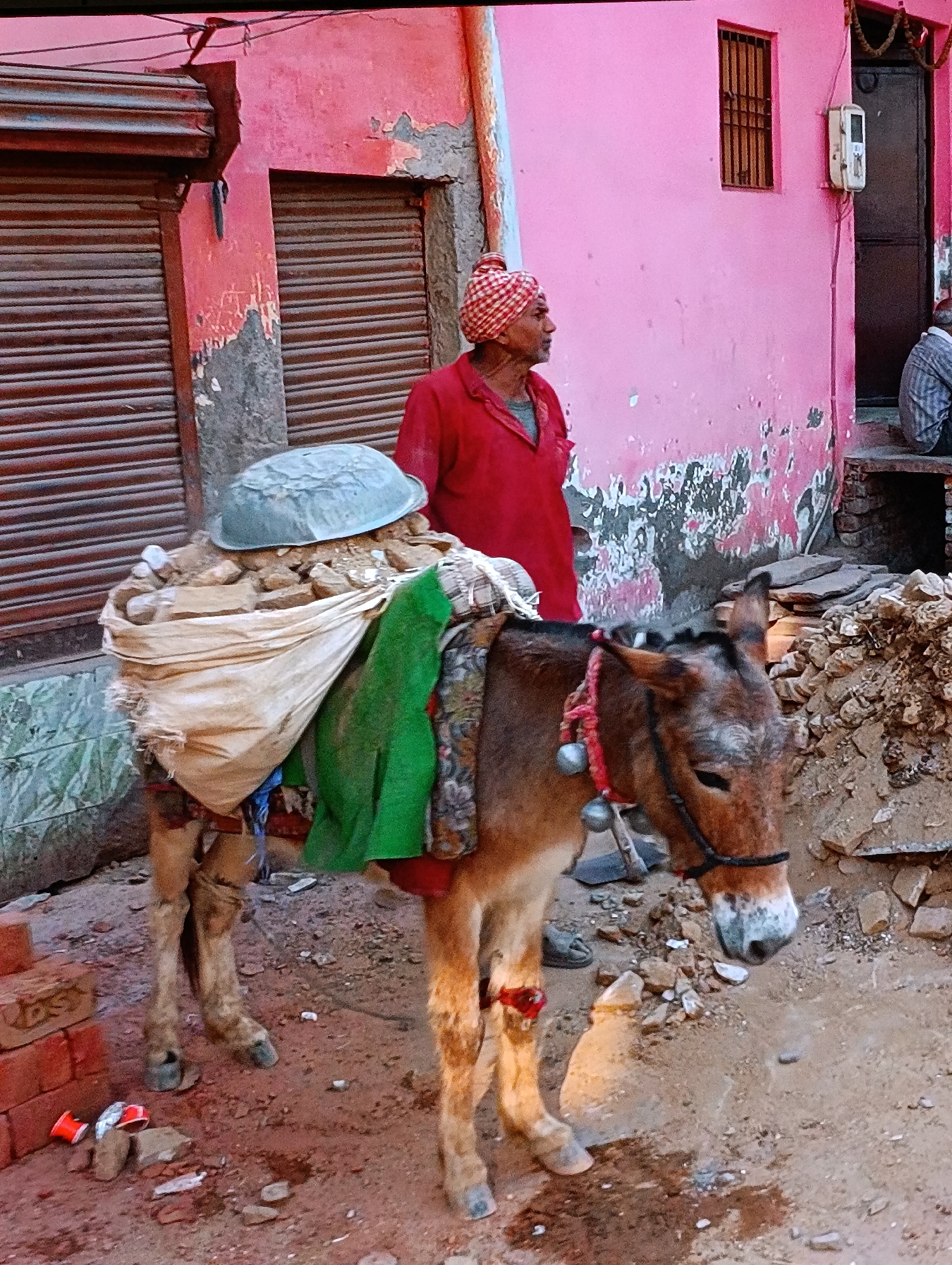 The image shows a donkey laden with a heavy load, standing on a dirt ground with debris scattered around. A man wearing a red shirt and a checkered head covering is standing near the donkey, looking towards the right side of the frame. The donkey is carrying a large sack filled with rocks or debris, topped with a metal bowl. The donkey has a bridle and bells around its neck. In the background, there are buildings with pink facades and closed metal shutters. A small barred window and an electrical meter box are visible on the pink building. The location appears to be an urban area, possibly a street or construction site, in Unknown, Unknown.