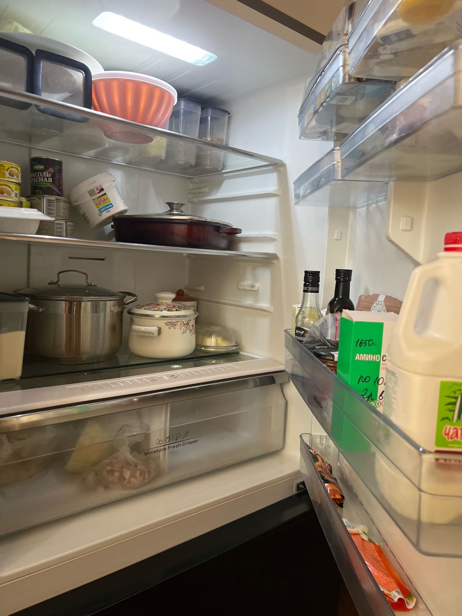 The image shows the interior of an open refrigerator. The refrigerator is white and appears to be a standard household appliance. On the top shelf, there is a large orange bowl and several clear plastic containers filled with what appears to be dry goods. Below that, on the next shelf, are several cans, a tub of sour cream, and a large pot with a lid. To the right of these items, there are bottles of oil and what looks like a box of amino acids. The bottom shelf of the refrigerator contains a large stainless steel pot, a smaller decorative pot, and a glass container of butter. In the crisper drawer below, there are bags of frozen food. The overall environment is clean and organized, suggesting the refrigerator is well-maintained. The image is taken from a slightly elevated angle, looking down into the refrigerator. The lighting in the refrigerator is bright, emanating from an LED strip light at the top. There are no people visible in the image. The location is not specified in the image itself, and therefore, it cannot be definitively placed in Novogagatli, Russia.