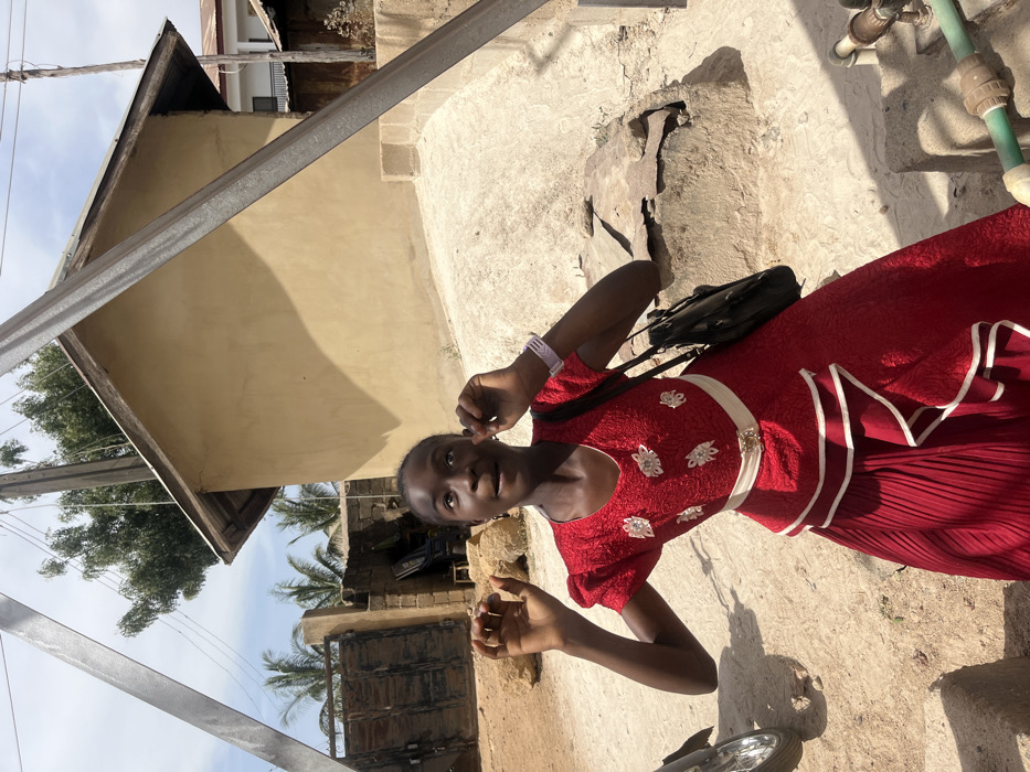 A young female stands outdoors on unpaved, sandy ground under a bright, sunny sky, in what appears to be a residential or semi-urban area in Jos, Nigeria. She is centrally positioned, wearing a textured red dress with short sleeves, white floral appliqués on the bodice, a white waist sash, and a pleated skirt with white-edged ruffles. A light purple wristwatch is visible on her left wrist, and a dark-colored handbag is slung over her left shoulder. Her dark hair is styled close to her head. Her hands are raised slightly with fingers splayed, and her head is tilted back, with her eyes looking upward and to the right, and her mouth slightly open.
In the background, to the subject's right, is a light brown, single-story building with a dark wooden eaves and a low-sloped roof. To her left, behind her, is a dark metal gate set within a concrete block wall. Several palm trees and other green foliage are visible further back. Two dark, diagonal metal beams, likely part of an overhead structure, traverse the upper portions of the frame. To the immediate right of the subject, a weathered concrete structure, possibly a wellhead, is present, showing exposed rebar and green PVC piping at the bottom right. A utility pole with overhead lines is visible to the left of the light brown building. A partial view of a motorcycle's front wheel and fender is visible on the far left. Shadows are cast on the ground and the building.