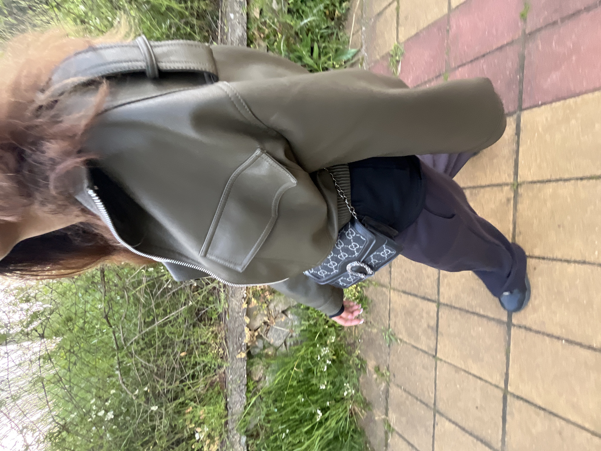 A person is standing on a sidewalk composed of rectangular tiles, some pink and some yellow. A green leather jacket with a buckled belt and front pockets is visible, along with a black purse featuring a silver design and a chain strap. The person is wearing dark gray pants and dark shoes. A metal fence is next to vegetation, which is on the left of the scene. The location is in a city park and not the city of Zlatograd, Bulgaria.