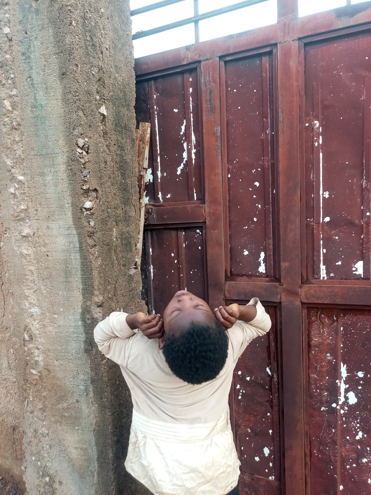 In Tempure, Nigeria, a young person stands in front of a weathered, red metal gate, their head tilted back and eyes closed. The gate is divided into panels, with peeling paint revealing rust beneath. To the left of the gate is a textured, concrete wall. The person is wearing a light-colored long-sleeved shirt and a matching skirt or wrap. Their arms are raised, hands resting near their ears. The scene appears to be outdoors, possibly in an alleyway or an unkempt yard. The sky is visible at the top of the frame, a bright, overcast white.