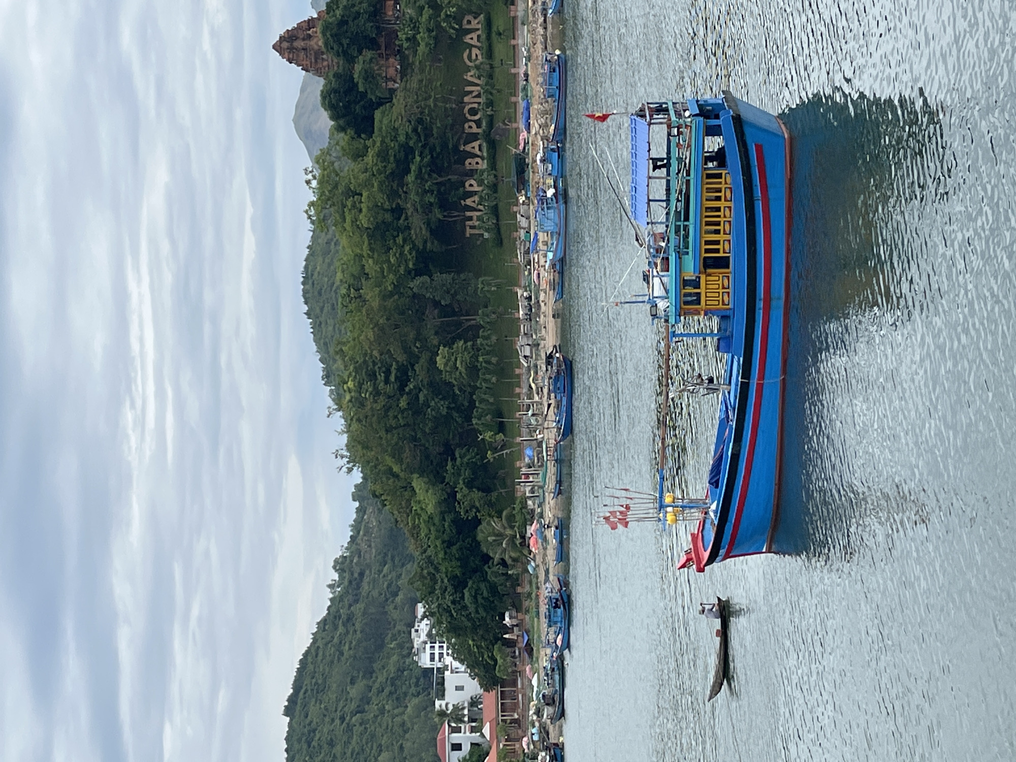 This image captures a waterfront scene in Nha Trang, Vietnam, likely near the Po Nagar Cham Towers, indicated by the prominent "THAP BA PONAGAR" sign on the hillside. In the foreground, a brightly colored blue and red fishing boat with yellow accents is moored on calm, rippling water. The boat has a multi-level structure with a blue roof and appears to be unoccupied or with minimal crew visible. To the left, a smaller, simple wooden boat is being rowed by a solitary figure wearing a hat.
In the middle ground, the shoreline is lined with numerous other fishing boats, some pulled ashore and others floating. A sandy beach is visible, with various materials, possibly fishing nets or tarps, spread out. Behind the beach, lush green trees and vegetation cover a hillside. Several buildings, including a white multi-story structure and smaller red-roofed buildings, are nestled among the trees.
Further in the background, the hillside continues, topped with more greenery and what appears to be a prominent, reddish-brown ancient tower, characteristic of the Po Nagar Cham Towers complex. The sky is overcast with soft, diffused light. The overall impression is a working harbor area with a backdrop of natural beauty and historical landmarks.