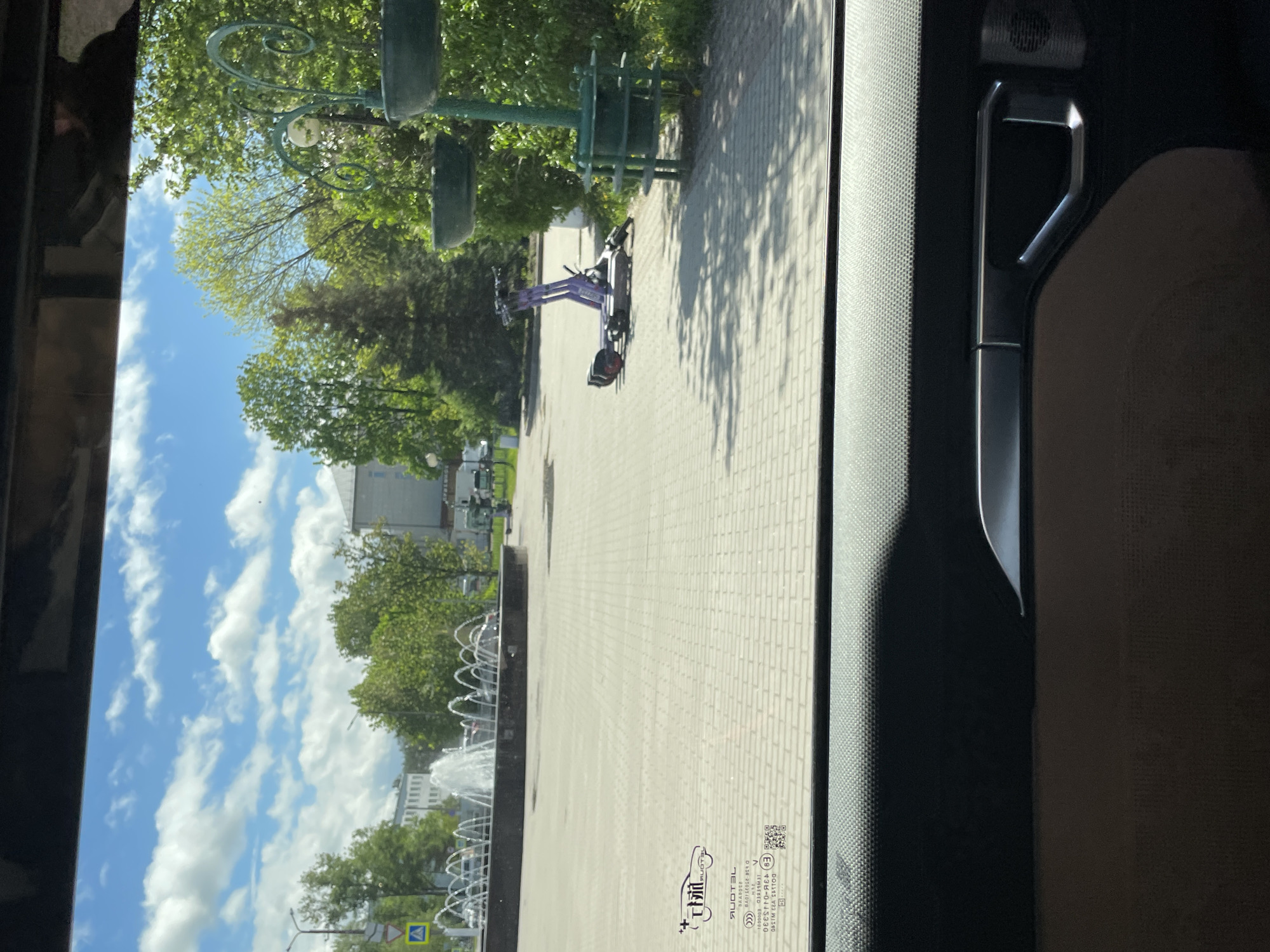 The image, taken from inside a vehicle, shows a view of a city street in Priozersk, Russia. Visible is a paved walkway leading to a fountain. An electric scooter lies on the ground to the right of the walkway, near a green lamppost with multiple tiers. A building can be seen in the background, along with trees and a blue sky with scattered clouds. The image is framed by the interior of the vehicle. The view is slightly tilted.