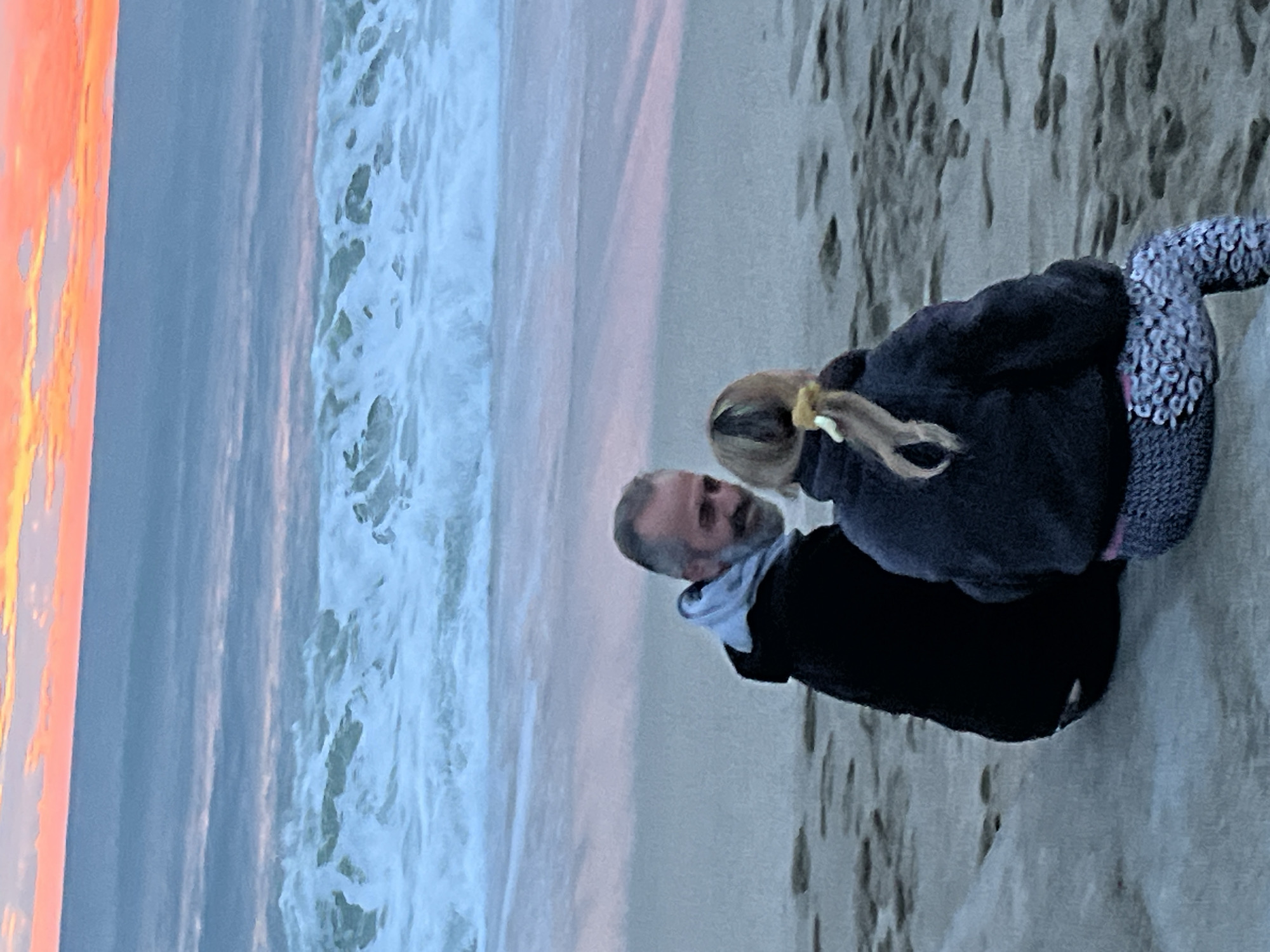 The image depicts an adult male and a child on a beach. The man has a beard and is wearing a dark jacket with a light-colored scarf. The child, partially visible, has blonde hair and is wearing a dark top and patterned leggings. They are standing on a sandy beach near the shoreline, with waves visible in the background. The sky shows a sunset or sunrise with orange and pink hues. It is impossible to state with certainty that the image was taken in Alanya, Turkey, without additional information or location markers.