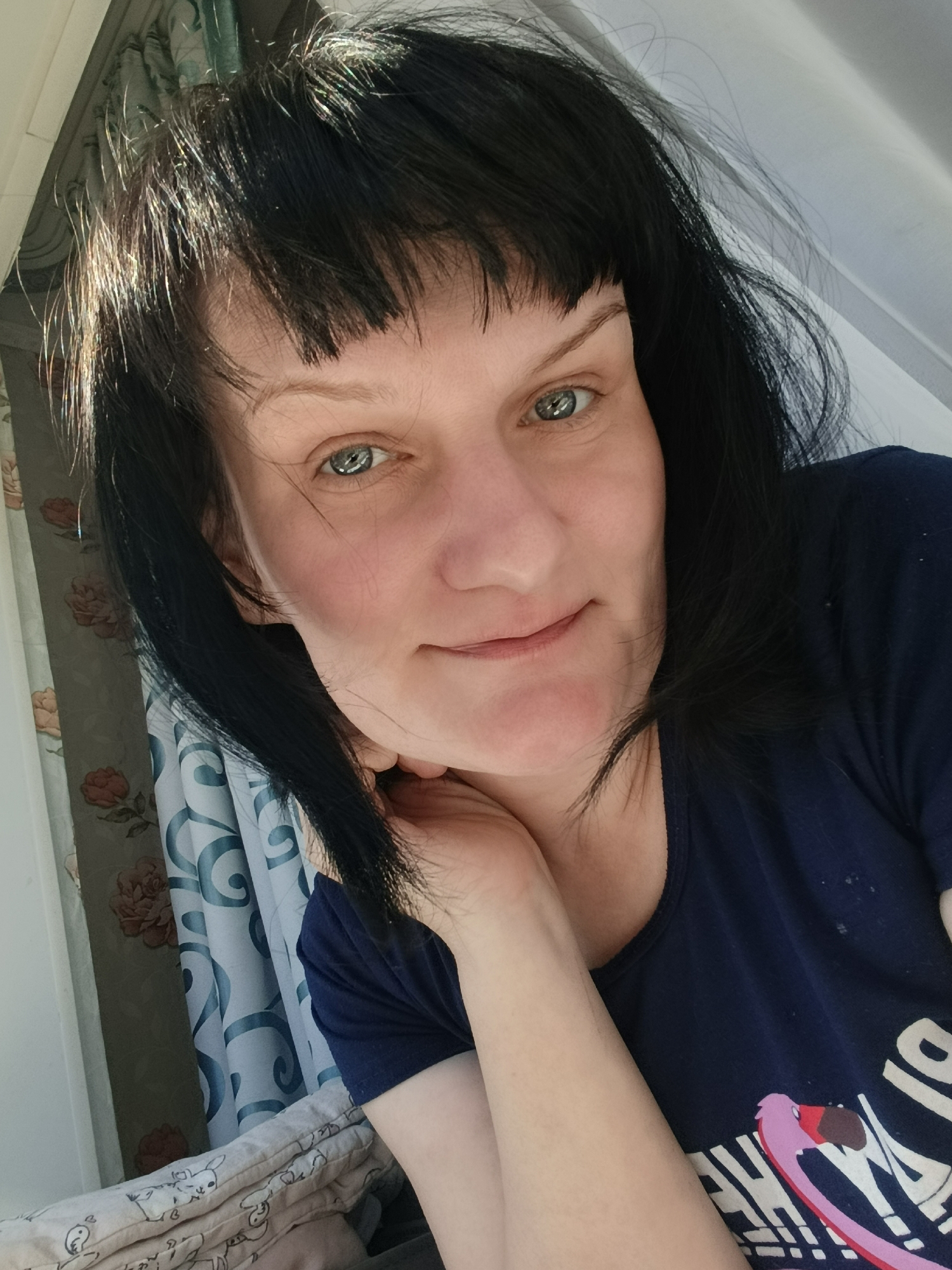 A close-up selfie depicts a woman with short, dark hair, including bangs, and fair skin. She is looking directly at the camera with a slight smile. Her left hand is raised to her cheek, with her fingers gently touching her face. She is wearing a dark blue t-shirt with a graphic print visible on the lower right side. The background includes a patterned curtain with floral motifs, a partially visible white frame structure and window. The image appears to have been taken indoors, possibly near a window, as suggested by the lighting. The image appears to have been taken in Фоки, Russia.