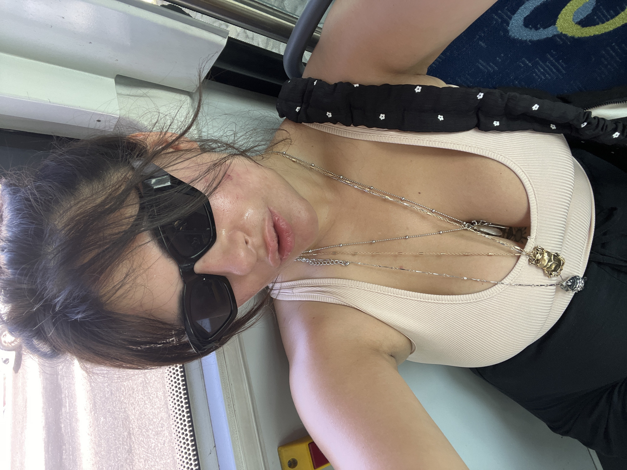 A woman with dark hair, wearing black sunglasses and a beige tank top, sits on a bus. She is looking at the camera and her lips are slightly parted. She is wearing two necklaces, one with a gold pendant and another with a silver pendant. The background of the image shows the interior of a bus, with a window and a seat visible. The image appears to have been taken on a sunny day, as evidenced by the bright lighting. The bus is likely in the city of Kiryat Bialik, Israel, as indicated in the prompt.