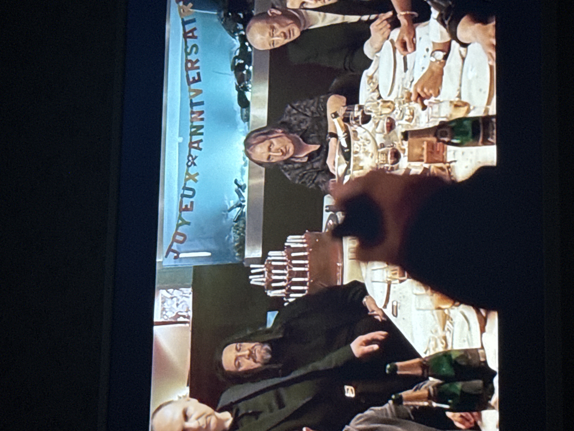 The image shows a group of people gathered around a long table set for a celebratory meal. Champagne bottles and glasses are prominent, suggesting a toast or special occasion. A banner reading "Joyeux Anniversaire" is visible in the background. The location is not discernible from the image to be Mytishchi, Russia. There are several people in the image around a table.