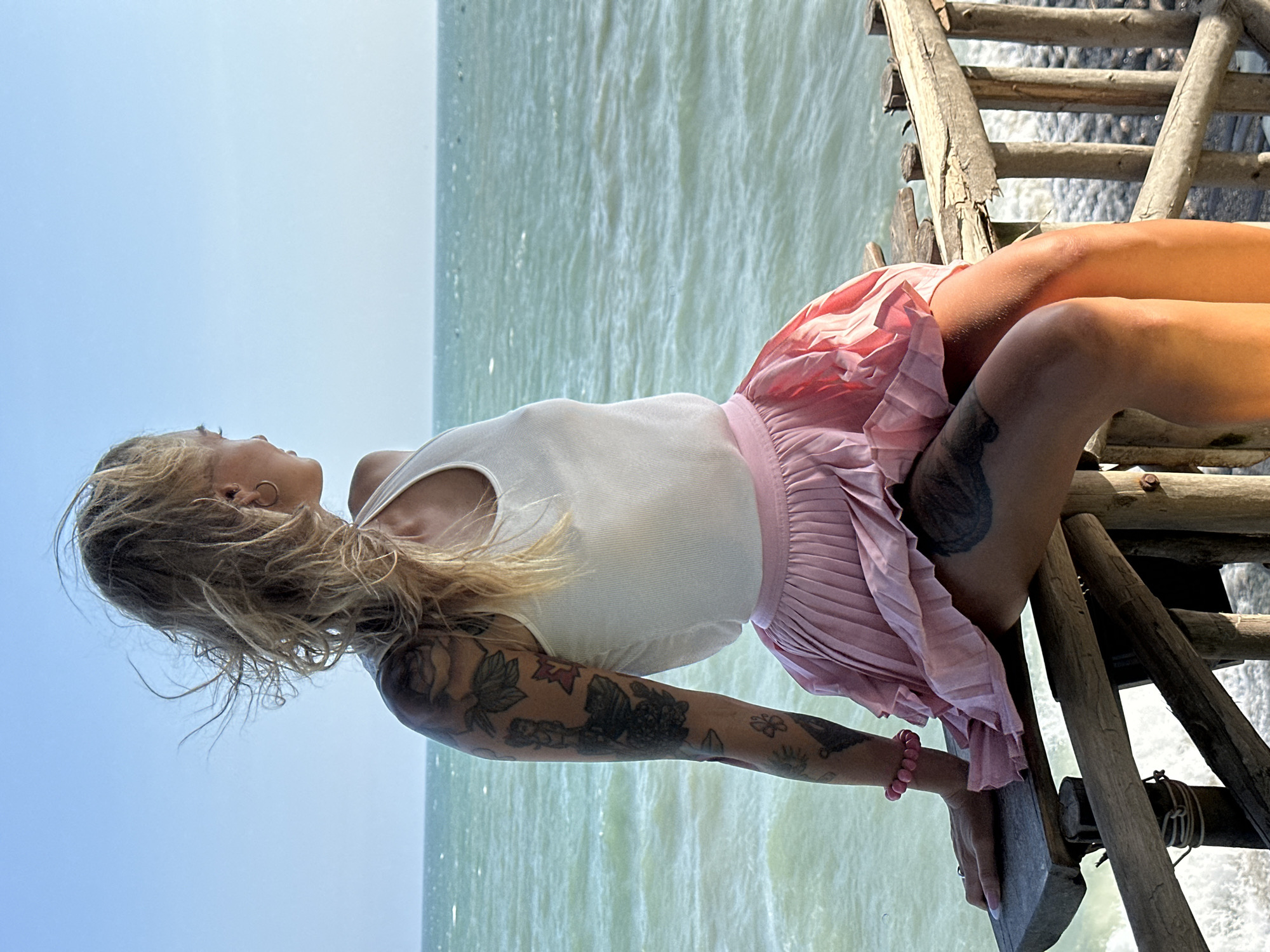 A young adult female with blonde wavy hair, wearing a white ribbed tank top and a pink pleated mini skirt, sits sideways on a weathered wooden railing, gazing out over a vast body of water under a clear blue sky. Her hair is windblown, and she wears small hoop earrings. Her left arm is extensively tattooed with various designs including floral motifs, and she has a pink beaded bracelet on her left wrist. Additional tattoos are visible on both thighs. Her left hand rests on the wooden surface. The water is choppy with visible waves and whitecaps, extending to the horizon. The scene is set in Phu Thuy Ward, Vietnam.