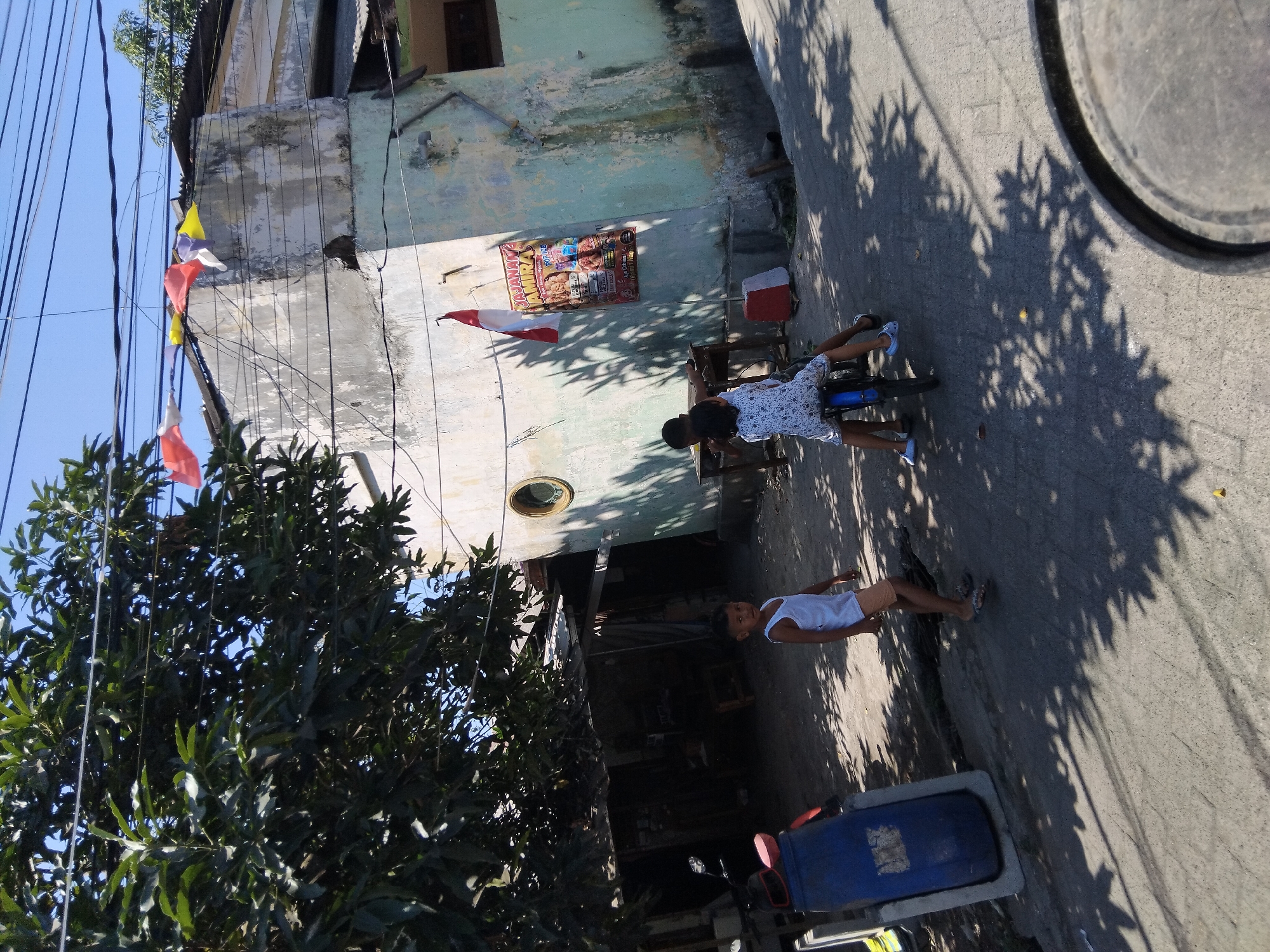 In Berbek, Indonesia, on a sunny day, two children are playing near a weathered building. A boy in a white tank top and peach shorts stands on a paved surface, looking towards the camera. In the background, another two children sit on a small motorcycle. The building behind them has an old, faded green and white exterior with a tin roof. A poster with red and white stripes, possibly advertising a business, is affixed to the wall. Flags are strung across wires above the building. A large blue barrel is positioned on the left side of the image. Lush green trees with dark leaves overhang the scene from the left, casting shadows on the ground along with the shadows of the tree leaves.