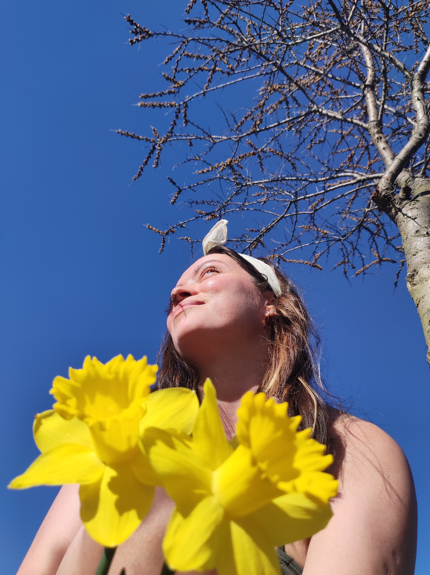 A low-angle, medium shot depicts a woman holding daffodils under a clear blue sky with a bare tree overhead. The woman, likely in her late twenties, has fair skin, light brown hair with blonde highlights, and wears a white headband with a bow. She looks upwards and slightly to the left, with a faint smile. The yellow daffodils fill the lower left quadrant of the image, partially obscuring her chest and shoulder. Above her head is the sparse branching structure of a tree, devoid of leaves except for small brown buds, set against the deep blue sky. The tree trunk is on the right side of the image. The scene conveys a sense of spring and warm weather. There is no information available to locate the specific location in the city of Фоки, Russia.