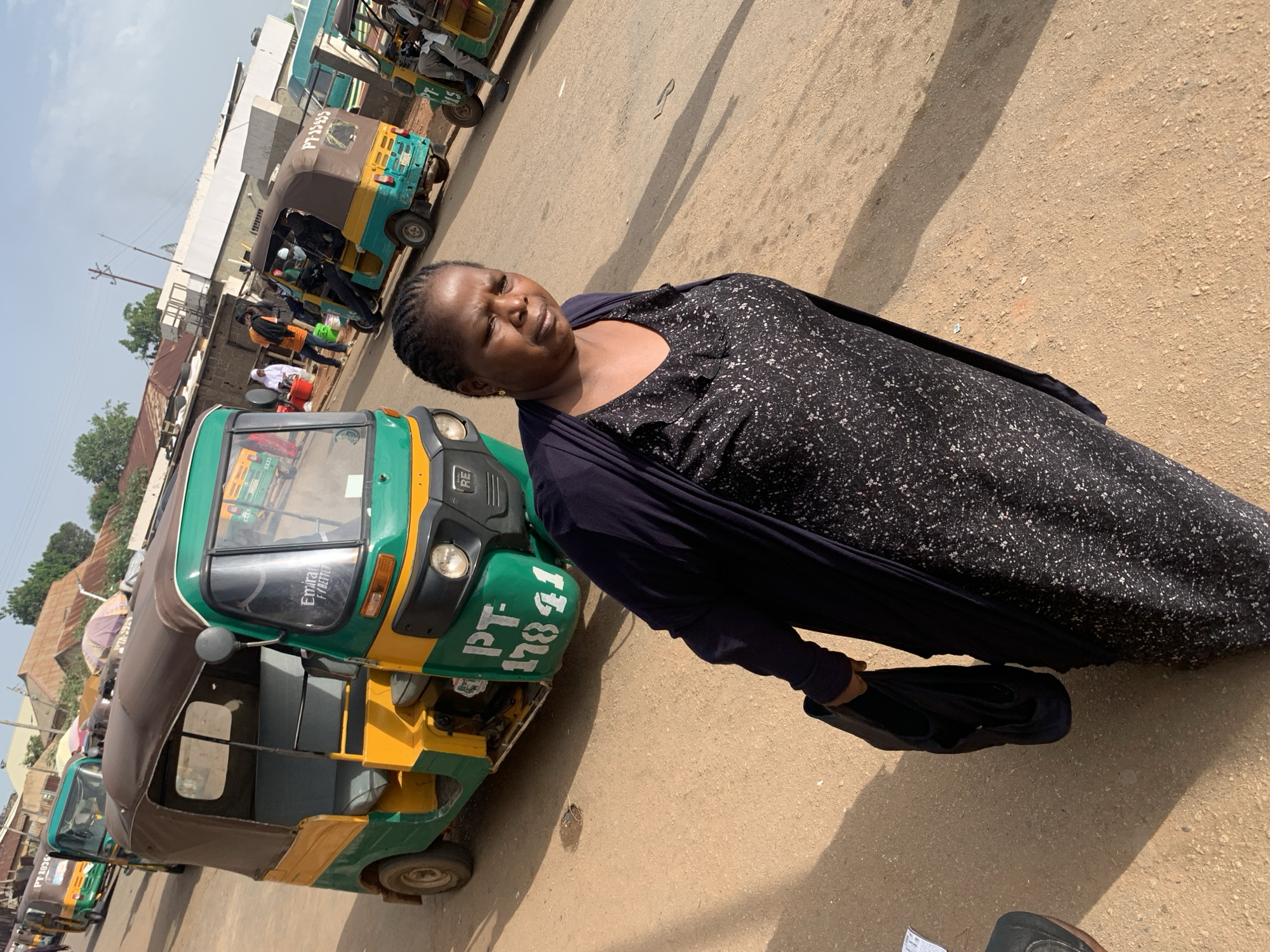 In the city of Gura Top, Nigeria, a woman is captured in a medium shot, standing on a dirt road. She is facing slightly to her right, her expression neutral, and she carries a black bag in her left hand. She is wearing a dark, patterned dress with a ruffled neckline and a long, dark cardigan. The background reveals a street scene with several tuk-tuks, some of which are colorful green and yellow. In the distance, buildings with corrugated metal roofs and some taller structures can be seen. The sky is visible above, suggesting it is daytime. The road surface is predominantly sandy and uneven, with some scattered debris. Other people are present in the background, some appearing to be passengers or drivers of the tuk-tuks, and one person is seen wearing a white garment and a headscarf. The overall impression is a typical street scene in a Nigerian town.