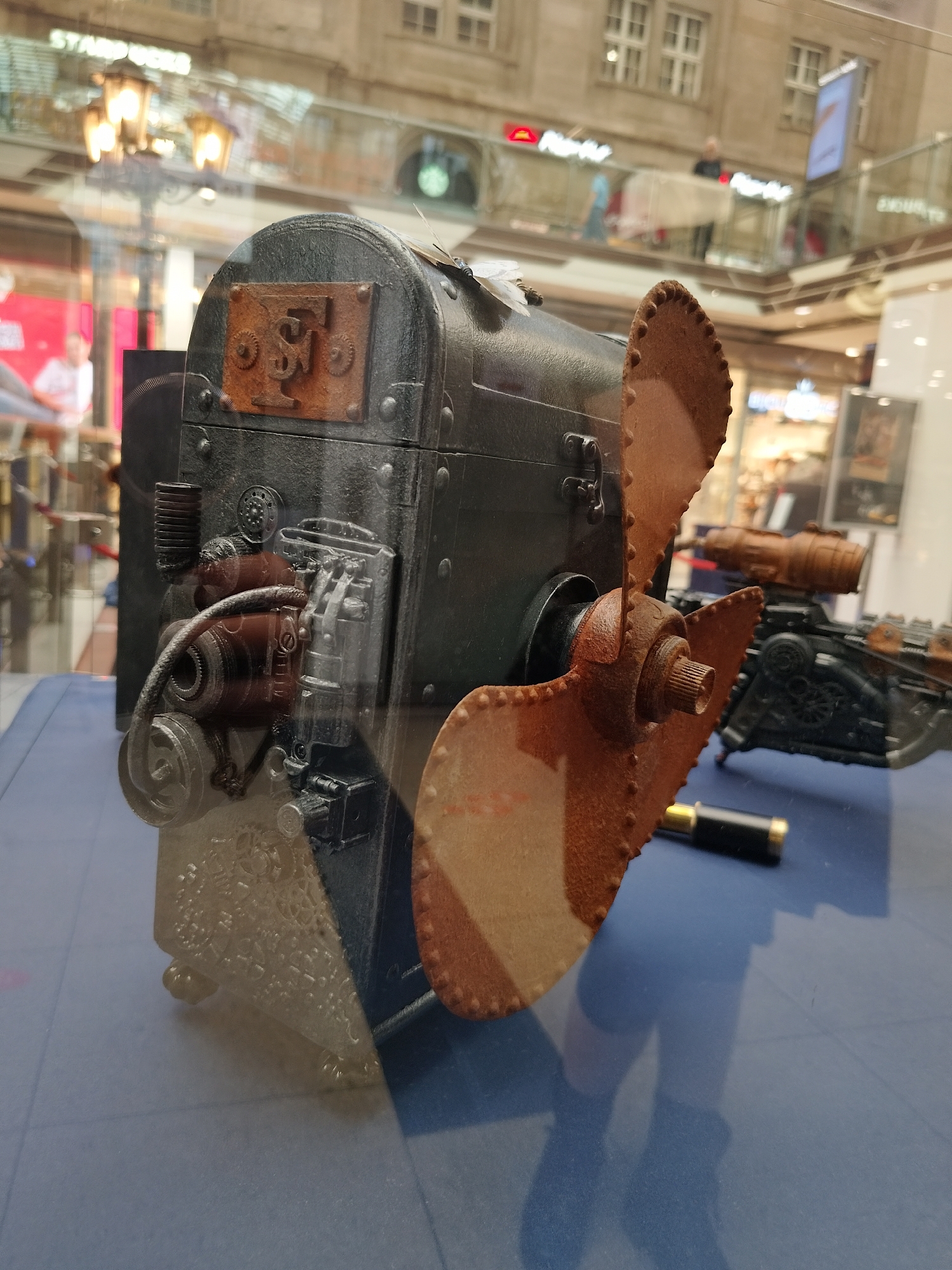 The image shows a steampunk-style model aircraft or boat, with a large brown propeller, displayed in a glass case. The model is primarily dark gray and brown. A small telescope or similar object lies next to it on the blue surface. The background reflection reveals a multi-story shopping mall interior with signs, street lamps, and shoppers visible on different levels. The location appears to be a shopping mall in Unknown, Unknown.