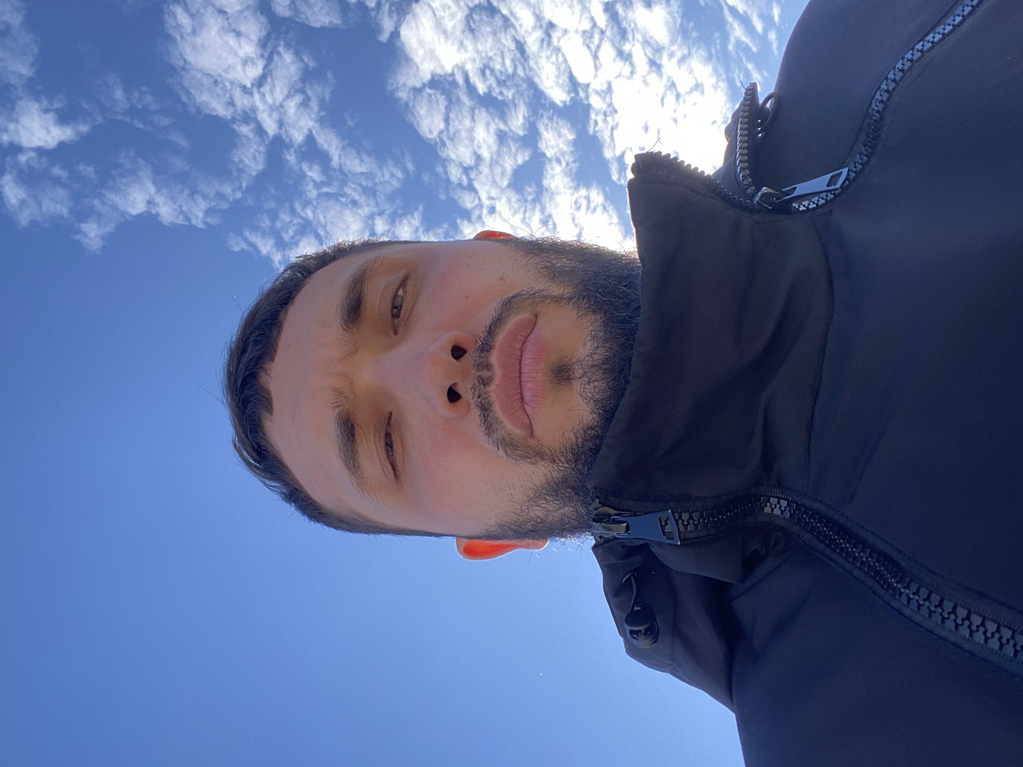 A man with a beard is looking directly at the camera from a low angle. He is wearing a dark jacket that is partially zipped. Behind him is a clear blue sky with some wispy white clouds. The location is unknown, and there is no evidence in the image to suggest it is Koskolovo, Russia.