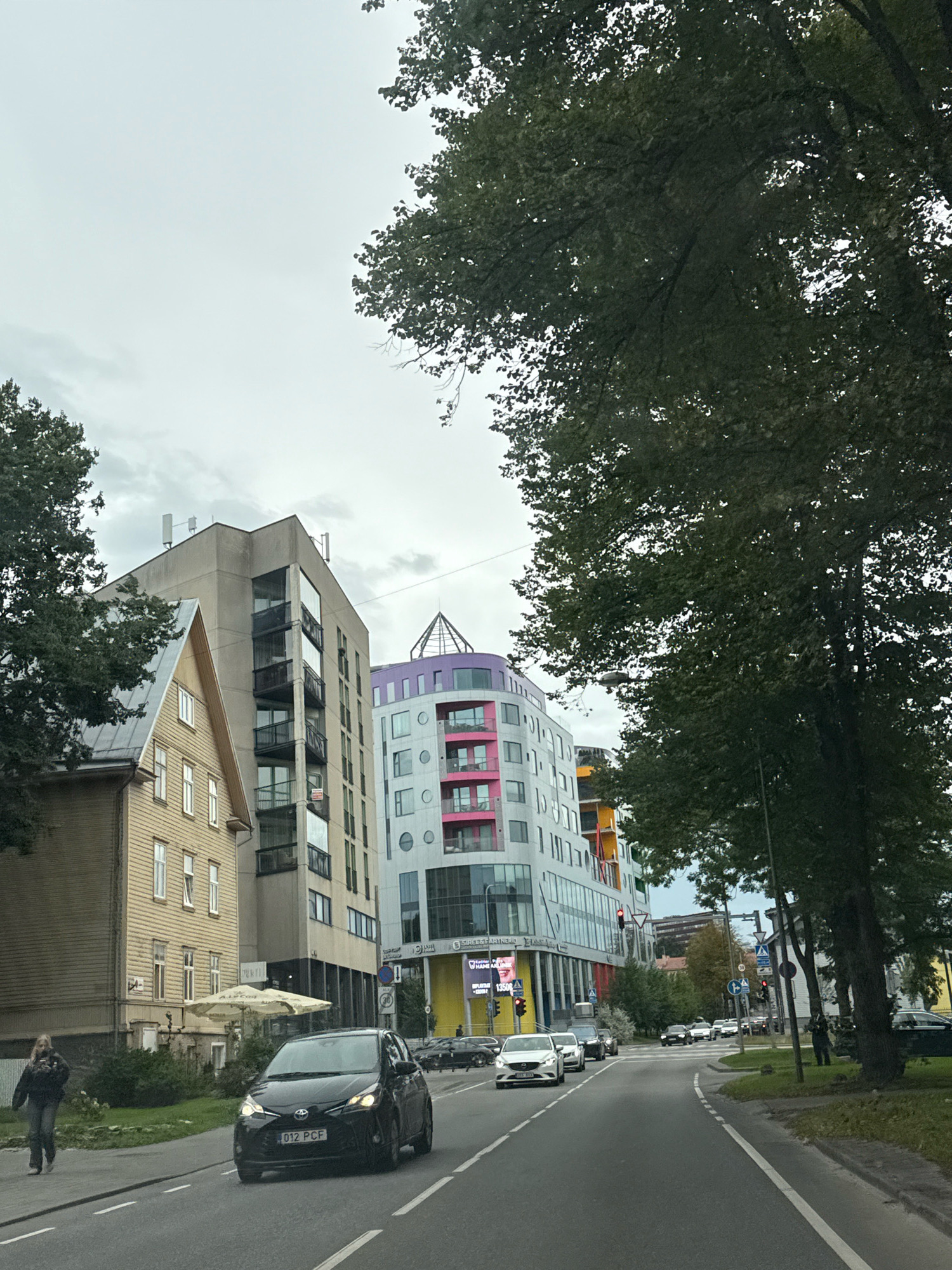 The image depicts a street scene in Tallinn, Estonia, on a cloudy day. In the foreground, a black Toyota Yaris with license plate "012 PCF" drives on the right side of the road. A woman in dark clothing walks on the sidewalk to the left of the car.
The street is lined with several buildings. On the left, a light-colored wooden house with a peaked roof stands next to a modern, multi-story concrete building with balconies. Further down the street, a rounded, colorful building features pink and grey sections and a pyramidal glass structure on top. Additional modern buildings with colorful accents are visible in the background, as well as trees with dark green foliage framing the right side of the image. Traffic lights and street signs are present along the road.