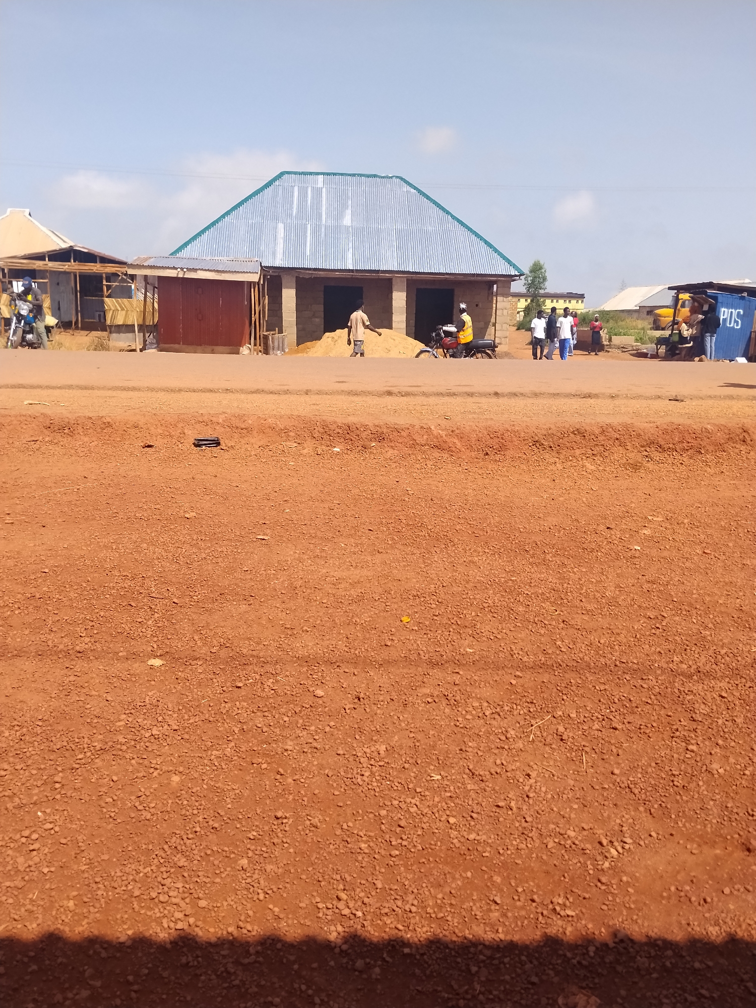 The image depicts a street scene in Durumi, Nigeria, characterized by a dusty, reddish-brown dirt road in the foreground. In the midground, several structures are visible, including a building with a corrugated metal roof, seemingly under construction or recently completed, with piles of sand outside. Adjacent to it is a smaller, older structure with a wooden and corrugated metal facade. A red motorcycle is parked in front of one of the buildings, with a person in a yellow vest standing nearby. Several other individuals are seen walking and gathered in small groups. Further to the right, a blue shipping container with "POS" written on it is visible, along with a yellow truck. The background shows more buildings and trees under a clear, bright sky. The overall impression is of a developing or semi-developed area with commercial activity.