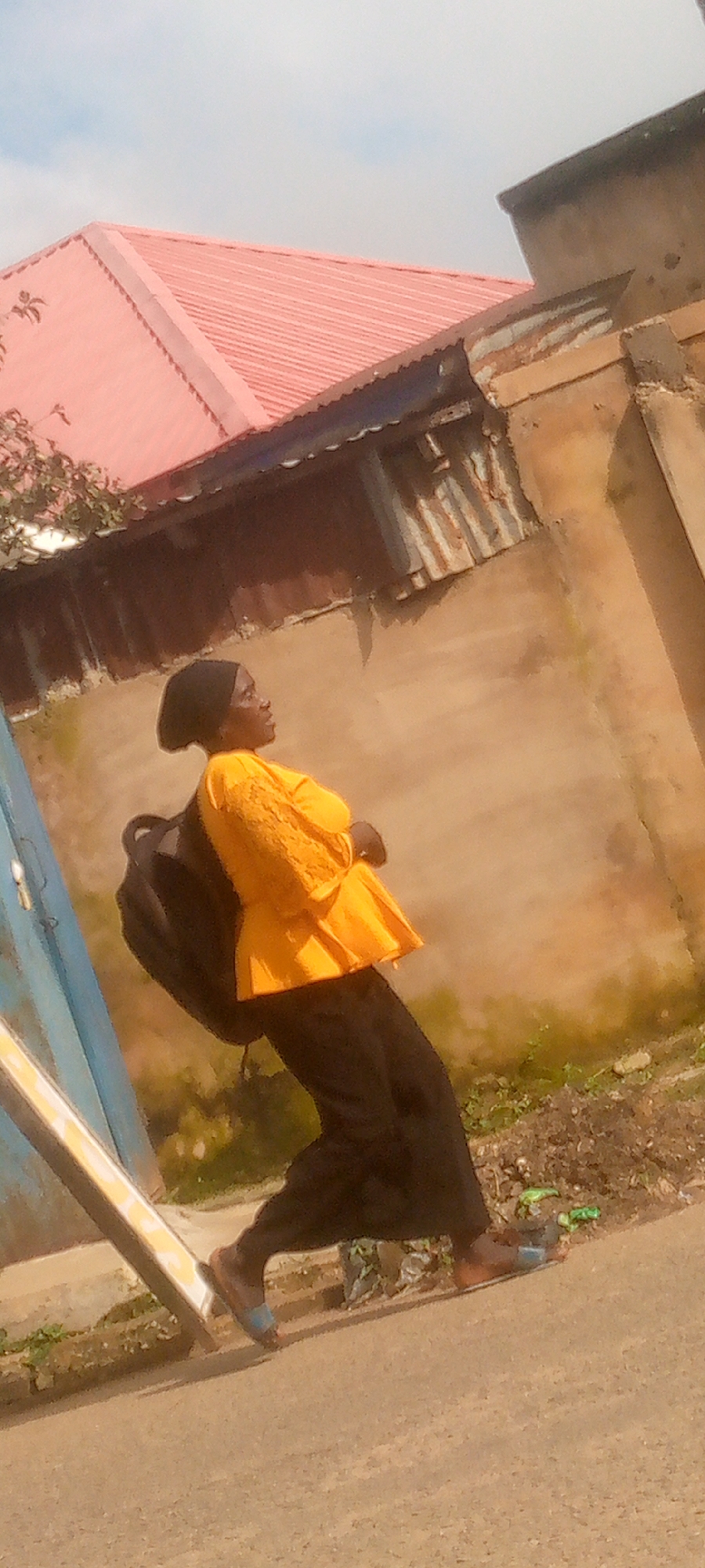 In an outdoor scene with a clear sky, a young woman wearing a black hijab and a bright orange, lace-detailed peplum top with wide black pants and blue slide sandals walks along a paved surface. She carries a black backpack over her right shoulder. To her left, a blue structure with a metal railing is visible. Behind her, a building with a light brown, textured wall and a corrugated metal roof section is present. A bright pink metal roof with a tiled pattern is visible above the building. The ground is littered with some green debris and white plastic. The image is taken from a slightly low angle.
