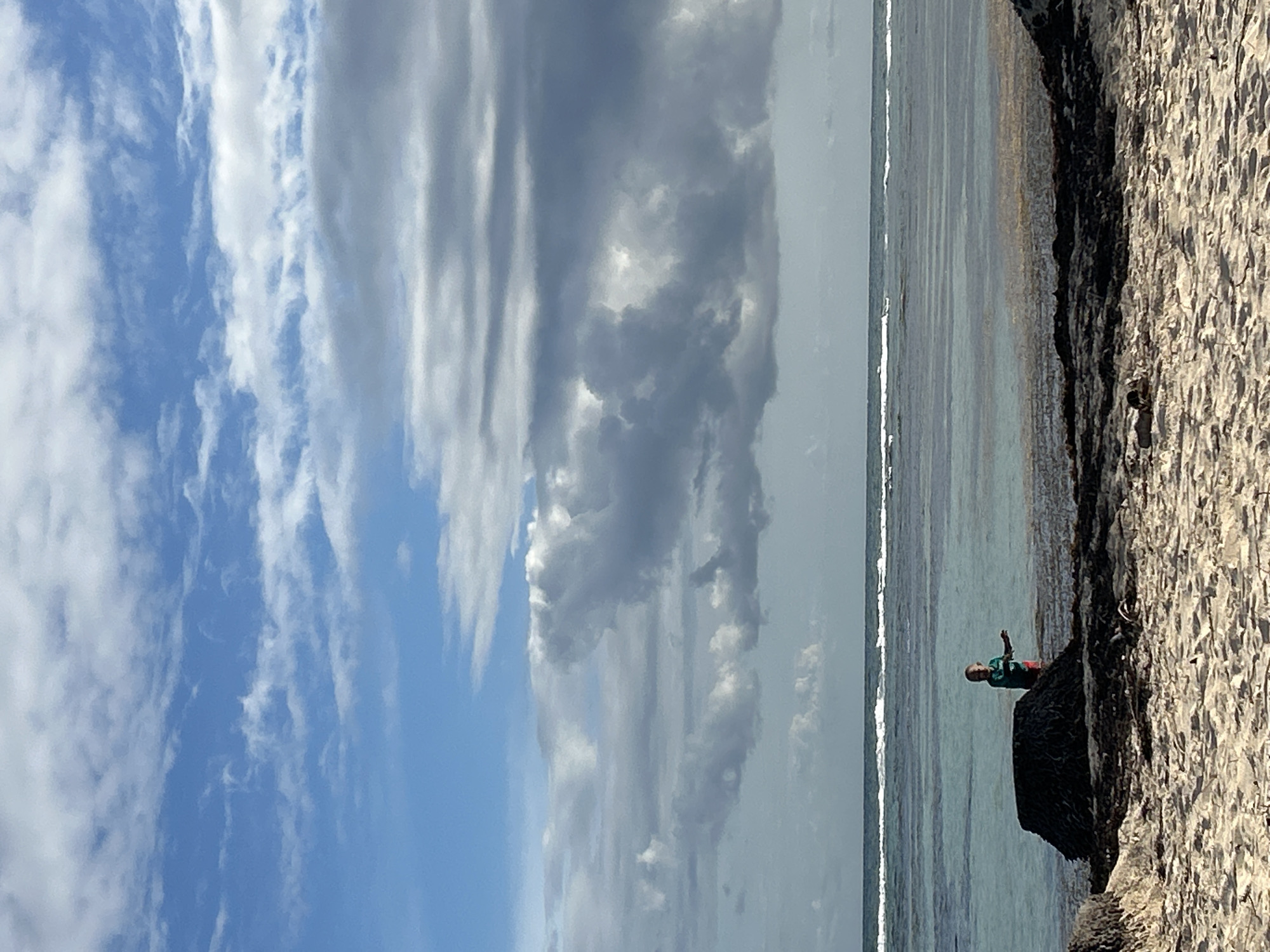 The photograph captures a coastal scene with a person standing on a dark rock formation near the water's edge. The location appears to be Sainte-Anne, France, based on the coastal geography and tropical atmosphere. The individual, presumably a child, is wearing a green shirt and red shorts, is standing near a dark rock that is on a beach of sandy white rock near the water's edge. The person is facing toward the sea and their right arm is raised into the air. The water is calm with faint small ripples breaking near the shore. The beach slopes gently into the sea. Behind the sea is a broad expanse of clouds, ranging from bright blue to grey. Some of the clouds are feathery and thin, while others are much darker and more dense. The sky and clouds take up the majority of the photograph, dwarfing the figure on the rock. The light is bright and diffused, suggesting a daytime setting with partial cloud cover. The image conveys a sense of solitude and tranquility.