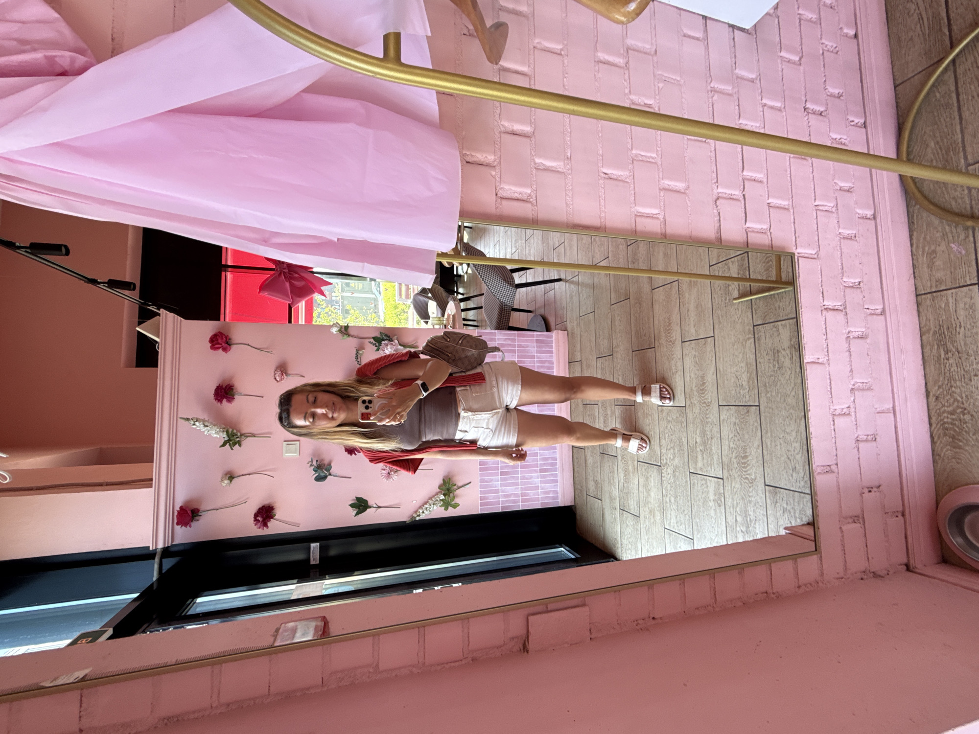 A young woman stands in front of a mirror in a room decorated with pink brick and floral accents. She is wearing a brown tank top, white shorts, and white sandals. She is holding a phone in her hand, taking a selfie. She has a small backpack slung over her left shoulder. The room appears to be a cafe or boutique, with tables and chairs visible in the background through a doorway. A pink fabric drapes from the ceiling on the right side of the image, and a gold clothing rack stands nearby. The overall impression is one of a trendy and feminine space.