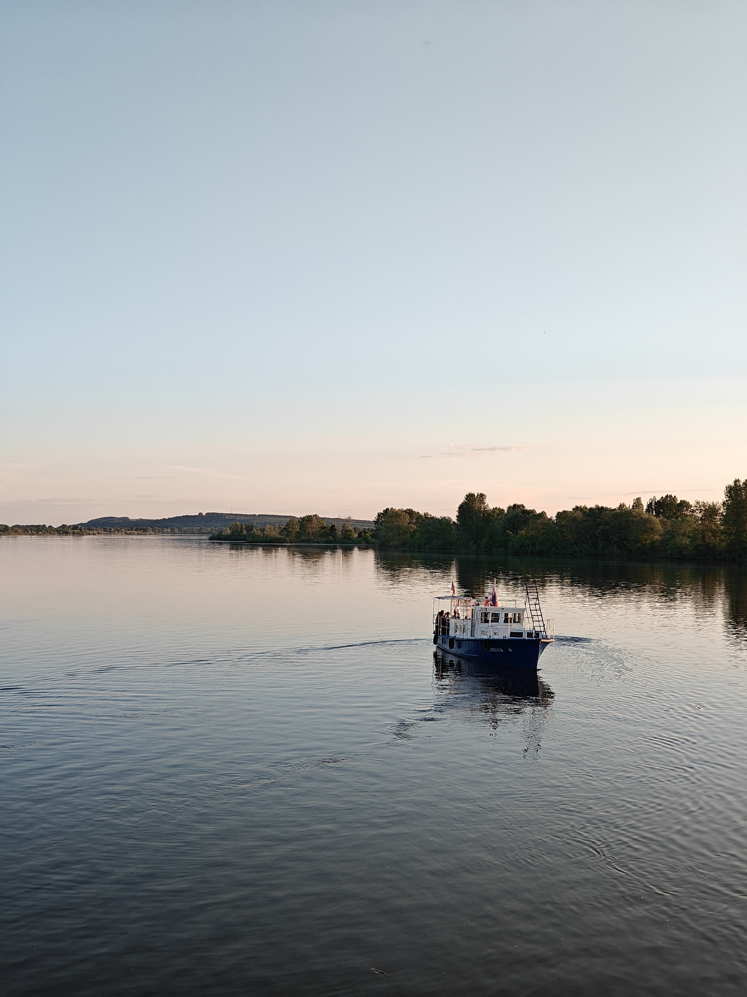 This image depicts a tranquil riverine scene at dusk or dawn, specifically located near Nikolo-Beryozovka, Russia, likely on the Kama River. The upper third of the frame shows a clear sky transitioning from a light, pale blue overhead to a soft pinkish-orange gradient at the horizon. Small, dark specks, possibly birds, are discernible in the upper sky.
The middle ground features a wide expanse of calm water, reflecting the sky's colors, with faint ripples extending across its surface. In the distance, a low-lying, densely forested landmass or shoreline stretches across the horizon, with a slightly elevated, treed hill visible on the far left. A smaller, tree-covered island or promontory is positioned closer to the left mid-ground.
A dark blue and white motorized pleasure boat is the primary subject in the lower-right quadrant, moving away from the viewer at a slight angle towards the right. It generates a visible wake trailing behind it, creating distinct ripples on the water. The boat possesses a white cabin with windows, a flat upper deck encircled by railings, and a ladder extending from the stern to the upper deck. A small flag, predominantly red and white, is visible near the stern on the upper deck. Multiple individuals, appearing as passengers, are standing on the upper deck, mostly on the port (left) side, observing the surroundings. The overall lighting is soft, suggesting a low sun angle, contributing to a serene atmosphere.