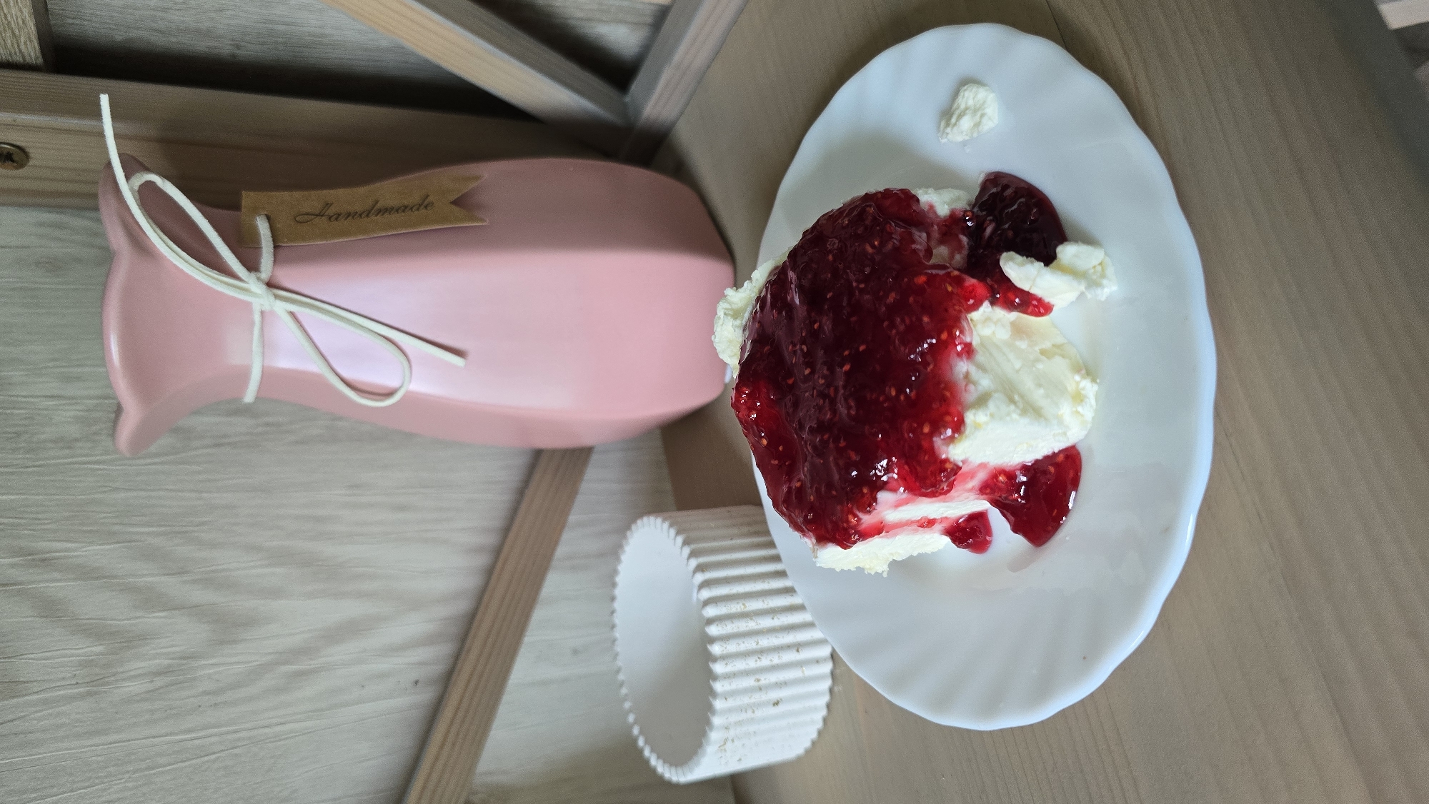 A slice of cream cake topped with raspberry jam sits on a white plate on a wooden table. To the left, a white ribbed cylindrical object is visible. In the background, a pink vase with a "Handmade" tag tied with white string stands on a wooden surface. The scene appears to be indoors, possibly in a home or cafe in Kyiv, Ukraine.