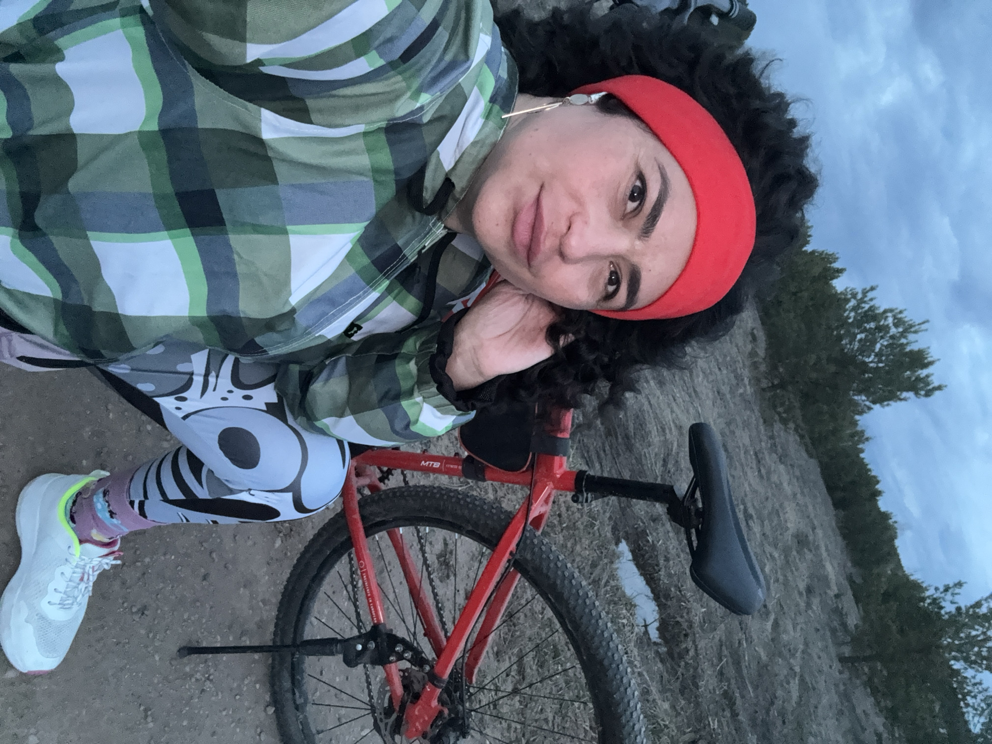 A person wearing a red headband, green plaid shirt, and patterned leggings poses with a red mountain bike in an outdoor setting. The background includes a dirt path, sparse vegetation, and a cloudy sky. The location is assumed to be near Кемуль, Russia.