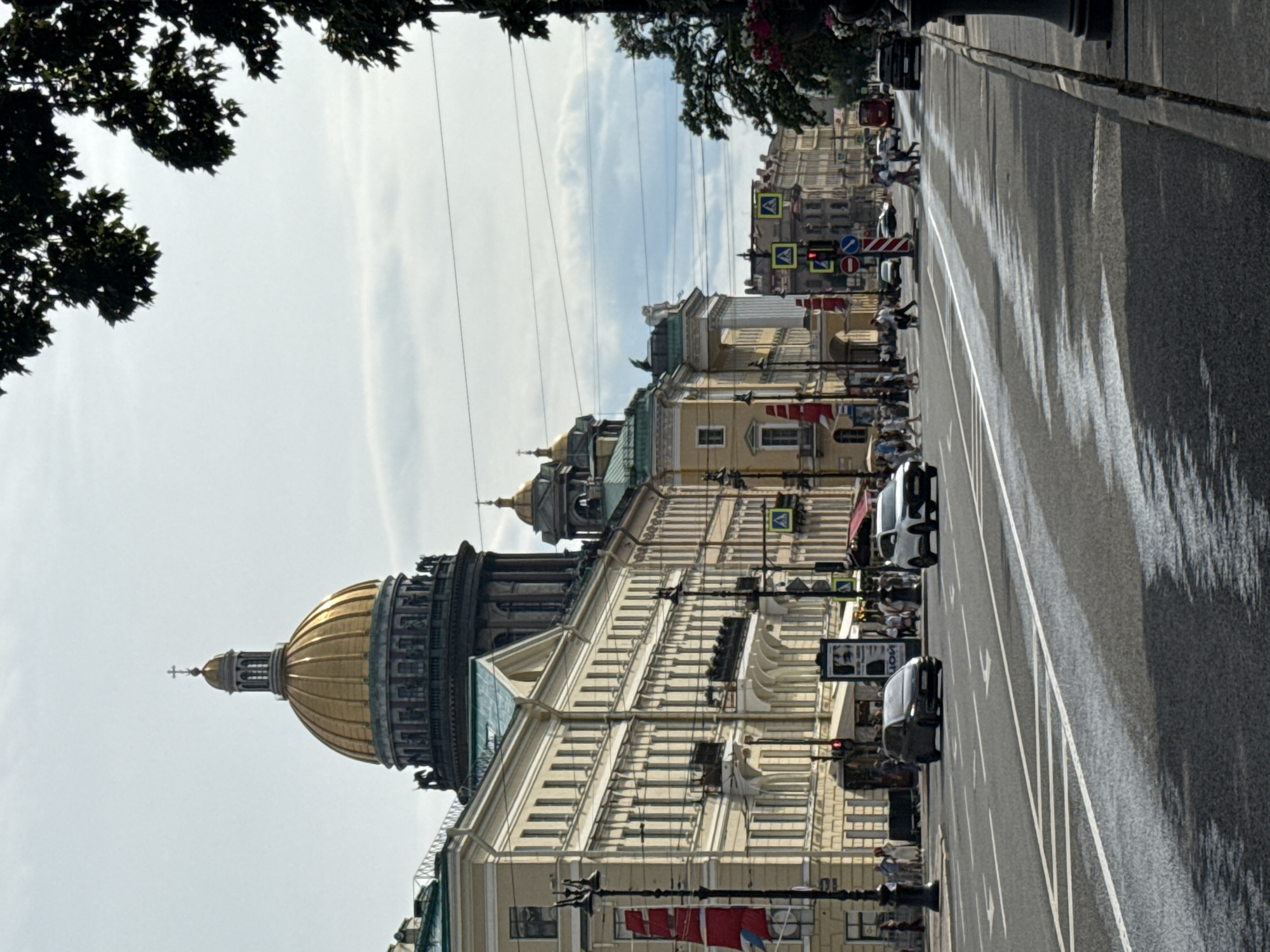 The image captures a street scene in Saint Petersburg, Russia, dominated by the grand architecture of a large building with a prominent golden dome, likely St. Isaac's Cathedral, visible on the left. The building is a pale yellow, with intricate classical detailing, multiple windows, and decorative sculptures.
The street itself is paved and has lane markings. Several cars are visible, including a white minivan and a dark-colored sedan. Pedestrians are walking on the sidewalk and crossing the street, indicating a busy urban environment. Traffic lights and pedestrian crossing signs are also present.
The sky is overcast with some lighter patches, suggesting it is daytime, possibly mid-morning or afternoon. The lighting casts shadows on the street, with the trees on the left side of the image creating a dark silhouette against the bright sky. The overall mood is one of everyday urban life, with the historical architecture providing a majestic backdrop.
There is some visible text on a sign in the middle right of the image, though it is partially obscured and difficult to read. Some red flags or banners are also hanging from a building. The perspective of the image is slightly tilted, adding a dynamic element to the composition.