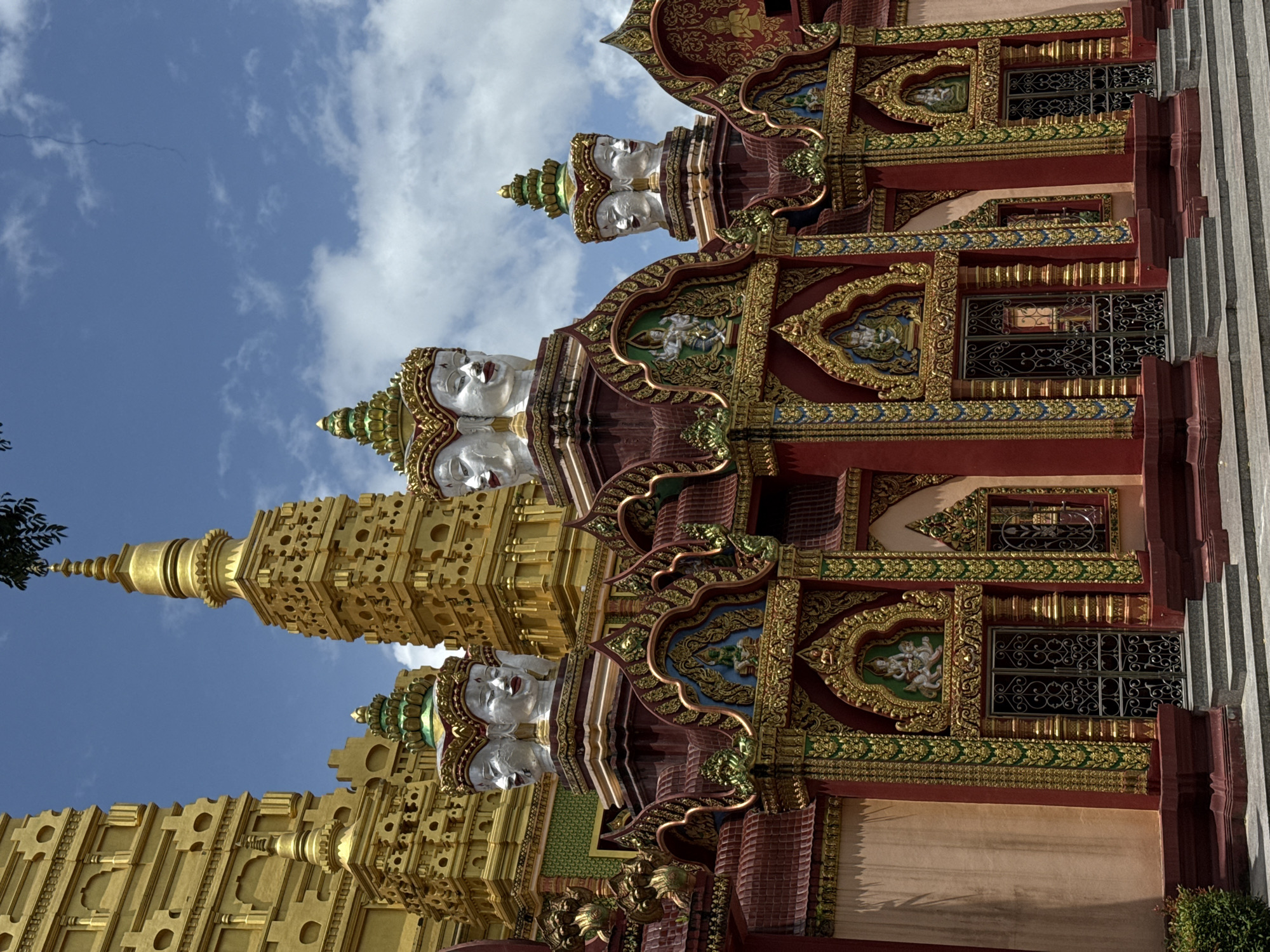 The image depicts the exterior of a temple in Na Nuea, Thailand. The temple features a striking facade with ornate architecture and religious iconography. Prominently displayed are several statues of multi-faced heads, with some appearing to have four faces each. These heads are white with red lips and are adorned with golden headdresses.
Behind the facade, tall, golden stupas rise into a bright blue sky dotted with white clouds. The lower section of the temple is painted in rich colors, predominantly red and a muted pinkish-orange, with intricate gold and green detailing. Arched doorways with decorative metal gates are visible, and above each doorway, there are recessed niches containing figures. Steps lead up to the entrances. The overall impression is one of grandeur and spiritual significance.