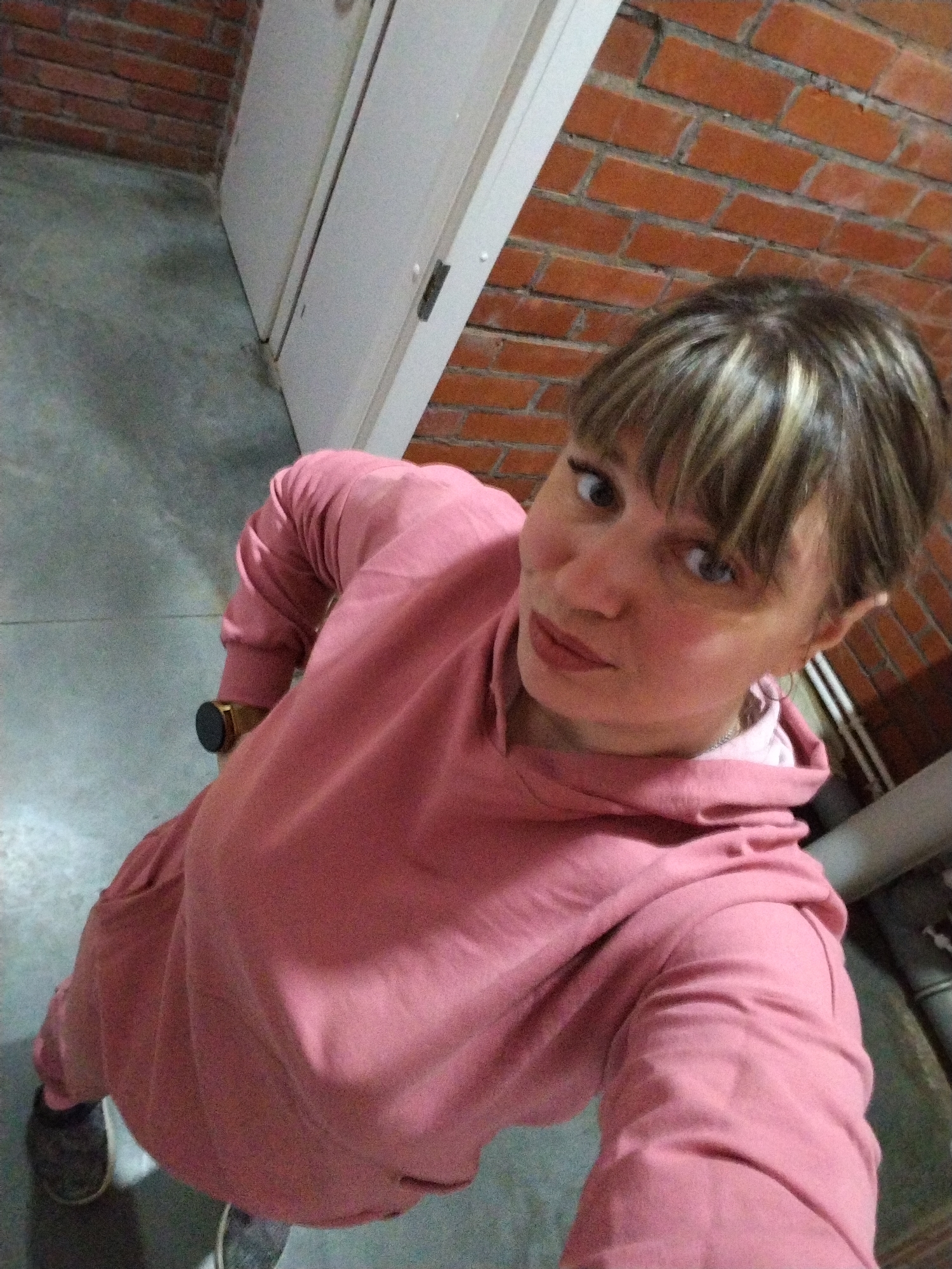 An adult woman takes a high-angle selfie indoors, likely in Rusanovo, Russia. She looks directly at the camera with a slight smirk. Her hair is short, dark brown with prominent blonde highlights, and styled with bangs. She has light-colored eyes, defined eyelashes, and her lips are painted with a subtle pink lipstick.
She is wearing a loose-fitting, dusty rose-pink tracksuit, consisting of a hoodie and matching pants. A round watch with a dark face and gold-colored casing is visible on her left wrist.
The background features a textured red brick wall to her right and behind her, suggesting an older building or industrial aesthetic. To her left, two white doors or door frames are visible. The floor is a plain, grey concrete surface. In the lower right corner, some white and grey pipes or conduits run along the brick wall. The overall lighting appears to be dim and artificial. There is no visible text in the image.