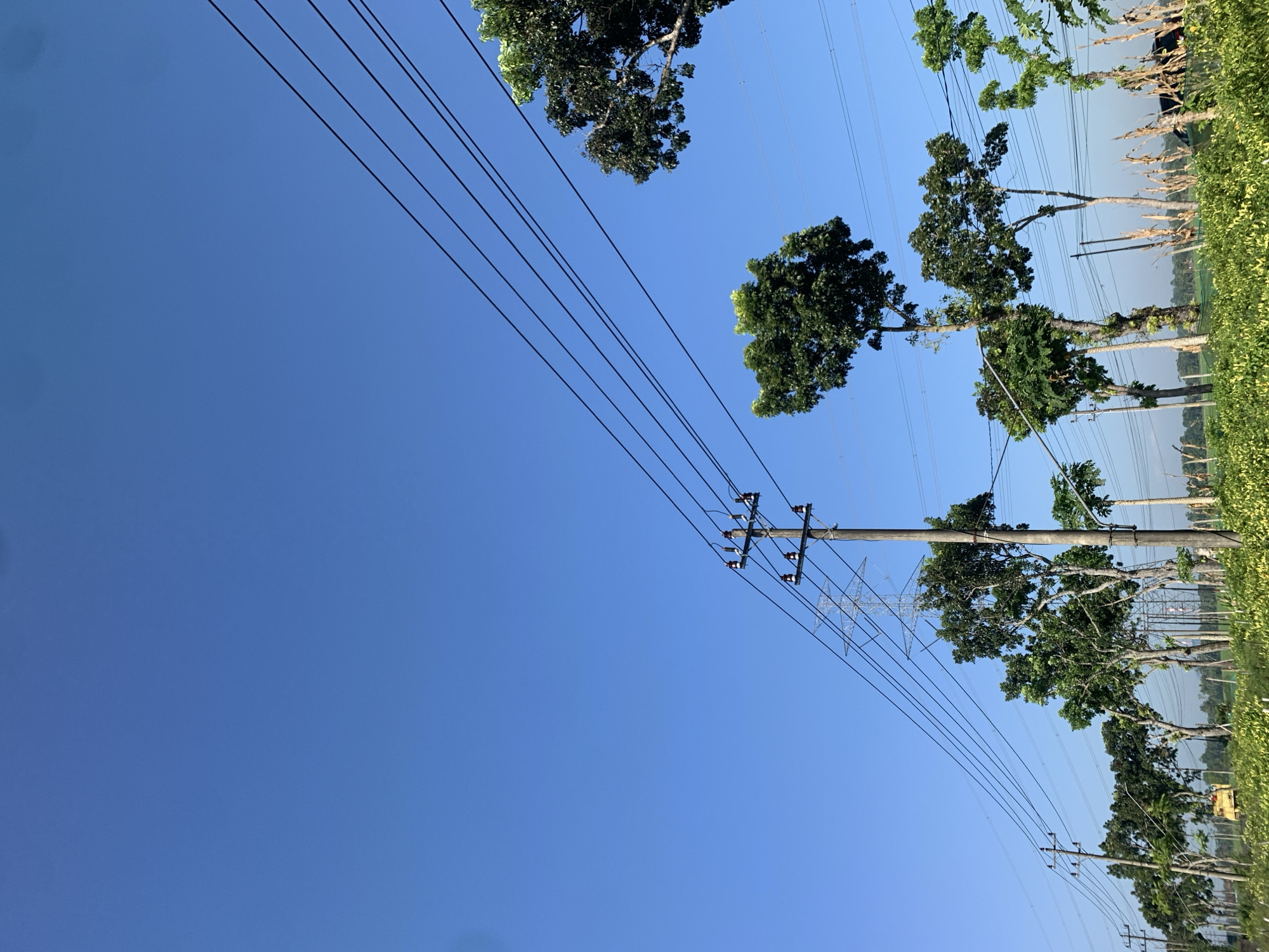 The image depicts a rural scene in Karangkemiri, Indonesia, under a clear blue sky. Multiple utility poles with power lines traverse the frame, stretching from the left foreground towards the upper right corner. A line of trees, varying in height and foliage, runs horizontally across the mid-ground, interspersed with more utility poles and distant, larger transmission towers. The foreground is dominated by a field of lush, green crops, possibly agriculture. In the far left of the frame, a yellow piece of agricultural machinery, likely a tractor, is visible. The overall impression is one of a working agricultural landscape integrated with essential infrastructure.