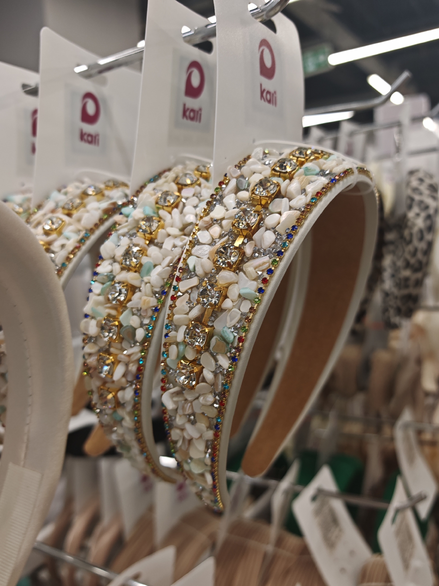 This is a close-up, eye-level shot of several ornate headbands displayed on a retail rack in what appears to be a store. The headbands are the primary subjects, characterized by their rich embellishments. They are covered in small, irregular pieces of what looks like white shell or stone, interspersed with colorful beads in shades of blue, red, green, and yellow. Prominently featured are clear, multifaceted gemstones set in gold-colored frames, adding a touch of sparkle. The headbands have a light brown interior.
Each headband is attached to a white cardboard tag, which has a pink logo and the word "kari" printed on it. These tags are suspended from a metal rack, suggesting a commercial display setting. The background is blurred, showing more merchandise on shelves and clothing racks, typical of a retail environment. The lighting suggests an indoor setting, possibly artificial, with no indication of time of day or weather. The focus is on the detailed craftsmanship of the headbands, highlighting their decorative elements. There is no discernible activity or event, as the image captures a static display. The visible text on the tags reads "kari". Given the context of Novy Urengoy, Russia, this would be a retail store in that location.