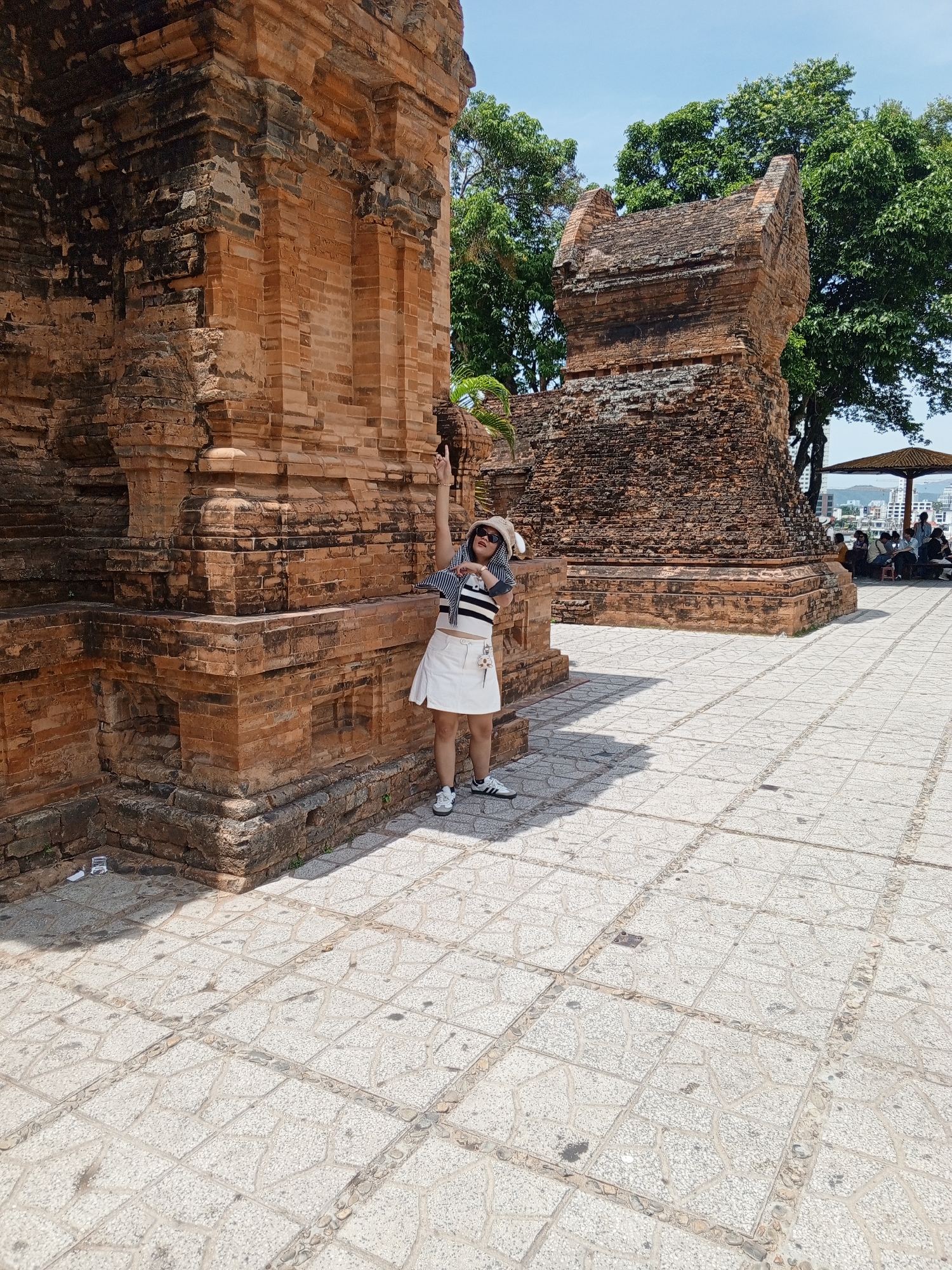 A woman stands in front of an ancient brick structure in Bac Nha Trang Ward, Vietnam. She is wearing a white skirt, a striped top, and a hat. The structure appears to be a temple or monument made of weathered red bricks, with a second, smaller structure visible in the background. The ground is paved with patterned tiles, and trees and a shaded seating area with people can be seen further in the distance. The sky is clear and blue.