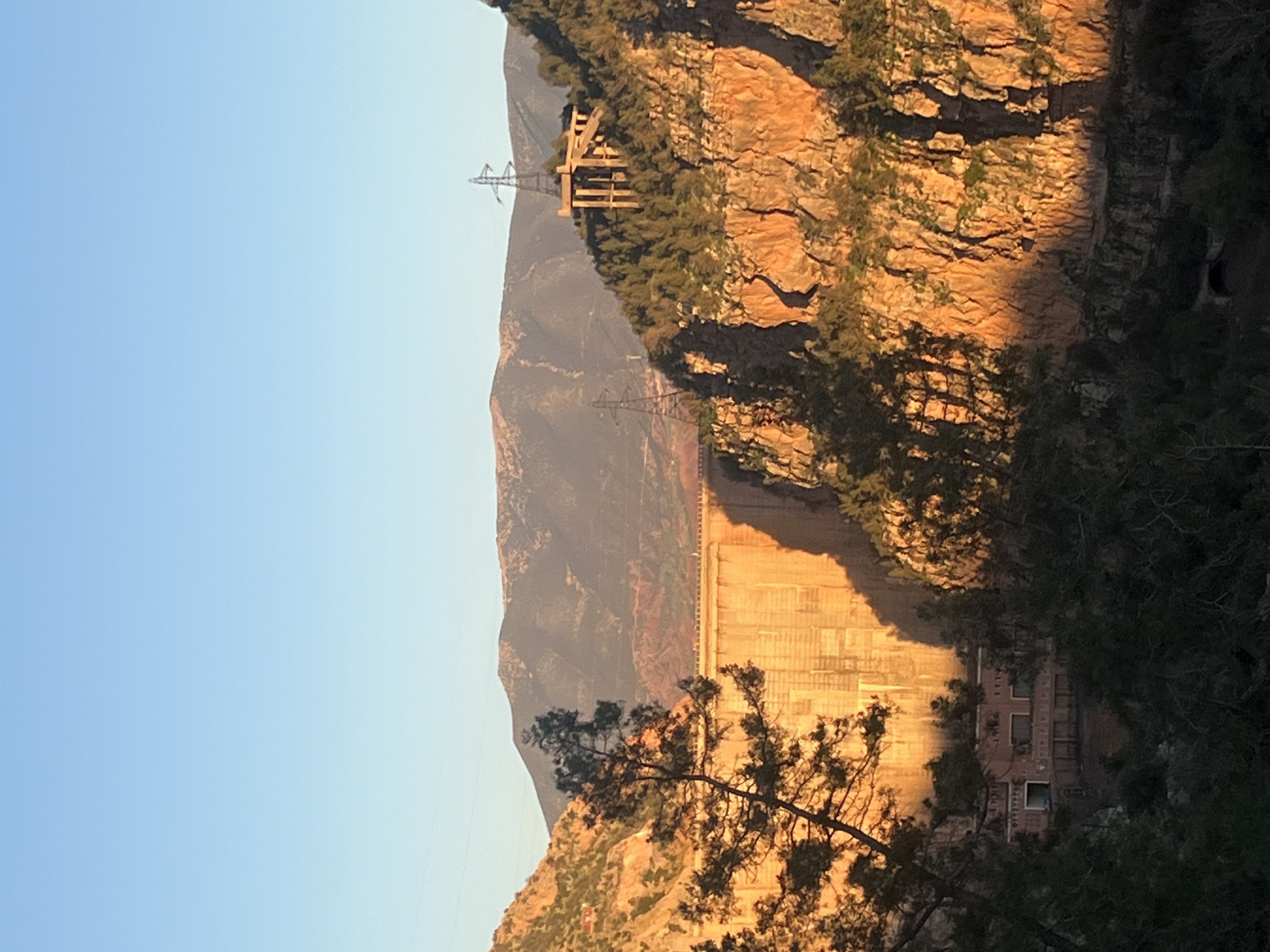The image shows the Bin El Ouidane Dam in Morocco. A large concrete dam wall stretches across the valley, casting a shadow. Power lines run across the mountaintops. The sky is clear and blue. Trees are growing on the sides of the mountain. A building is located at the bottom of the dam. The light suggests it is either sunrise or sunset.