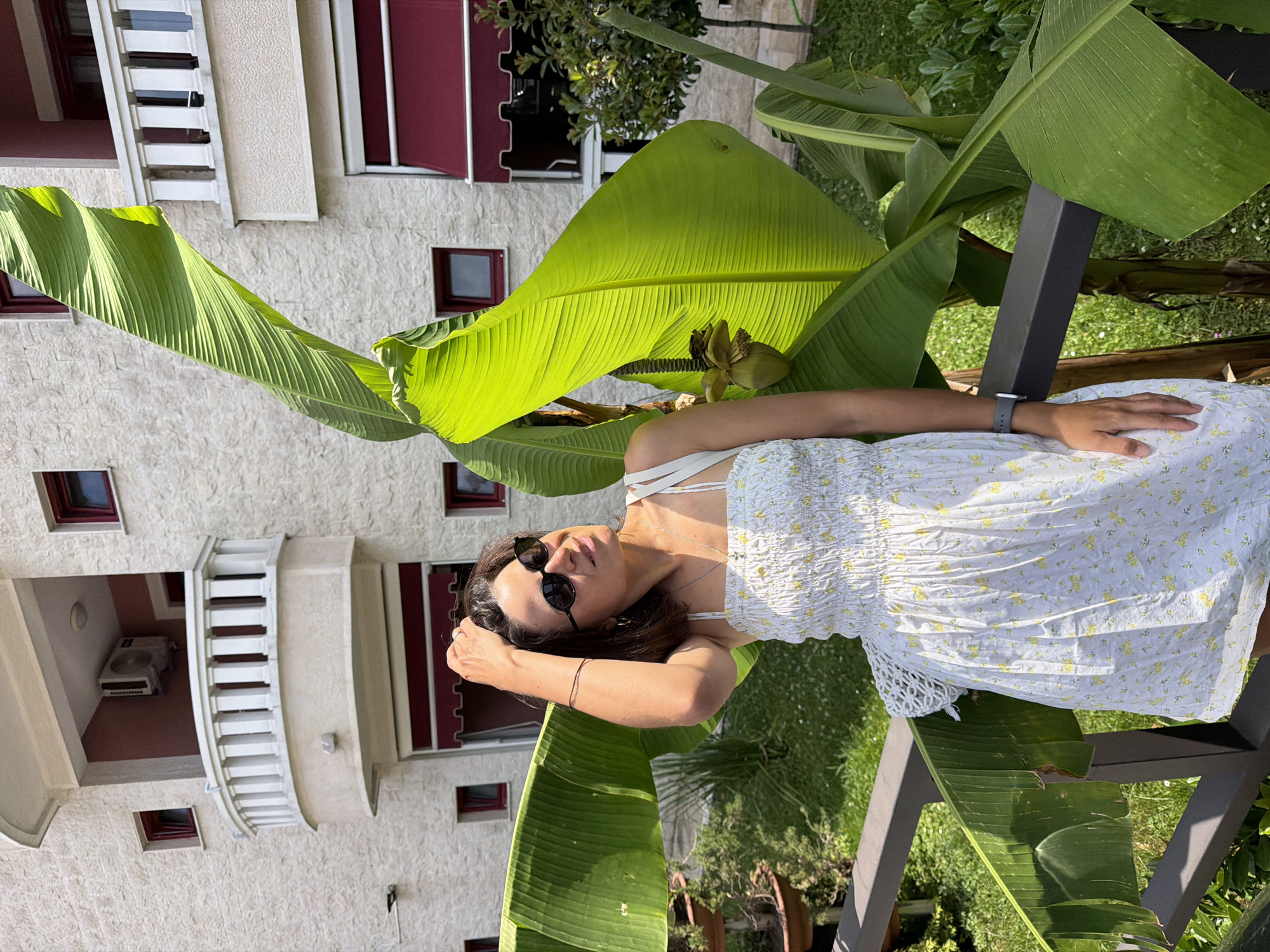 A woman stands outdoors in Budva, Montenegro, in front of a light-colored, textured building. She is wearing a white, floral-patterned dress with thin straps and sunglasses. Her right hand is raised, touching her hair. Large green banana leaves are prominent in the foreground and to her right, partially obscuring the building. A dark railing runs horizontally in front of her. The background shows a balcony with white railings and windows with red awnings on the building. Sunlight illuminates the scene, casting shadows and highlighting the vibrant green of the leaves.