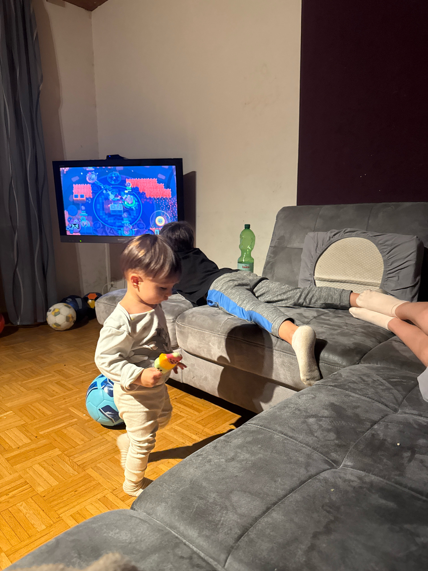 The image depicts an indoor scene, likely a living room, with a young child standing on a wooden floor and holding a toy. The child is wearing a light-colored long-sleeved shirt and cream-colored pants. A blue soccer ball is positioned near the child's feet. In the background, a television screen displays a colorful game or animation. A gray sofa is present, with two individuals, likely children, lying on it. One person on the sofa is wearing gray sweatpants with blue stripes and white socks, while another individual's legs are visible, wearing pink socks. A green plastic bottle stands on the sofa. Several soccer balls are scattered on the floor near the television. The environment appears to be a home setting. There is no information in the image to indicate the location is in Überherrn, Germany.
