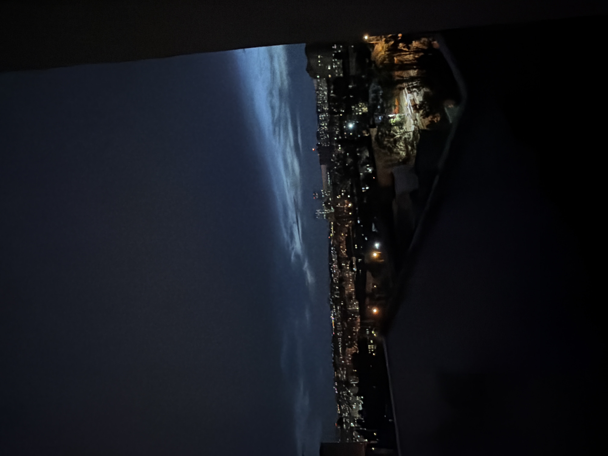 The image shows a nighttime view of a city, likely Kyiv, Ukraine, from a high vantage point. The sky is dark with some wispy clouds illuminated by faint, distant city lights. Below, the city is a sprawl of buildings with numerous lights indicating activity. The foreground is dominated by dark, shadowed areas, suggesting a balcony or rooftop view. The city lights create a warm glow against the dark backdrop of the night. The overall impression is one of a vast urban landscape at night, with the characteristic glow of a metropolitan area.