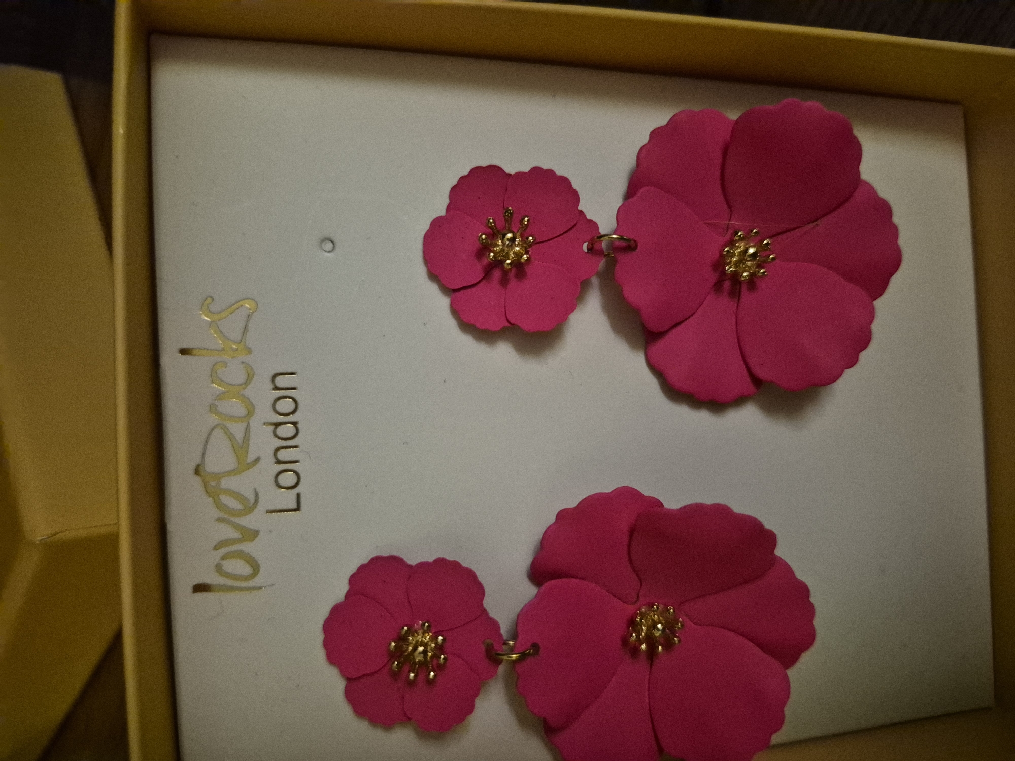 In a yellow box, a pair of hot pink floral earrings are displayed against a white background. The earrings are designed with multiple layers of petals, each with a gold bead center. The brand name "loveRocks London" is printed in gold foil above the earrings. The box appears to be part of a retail display.