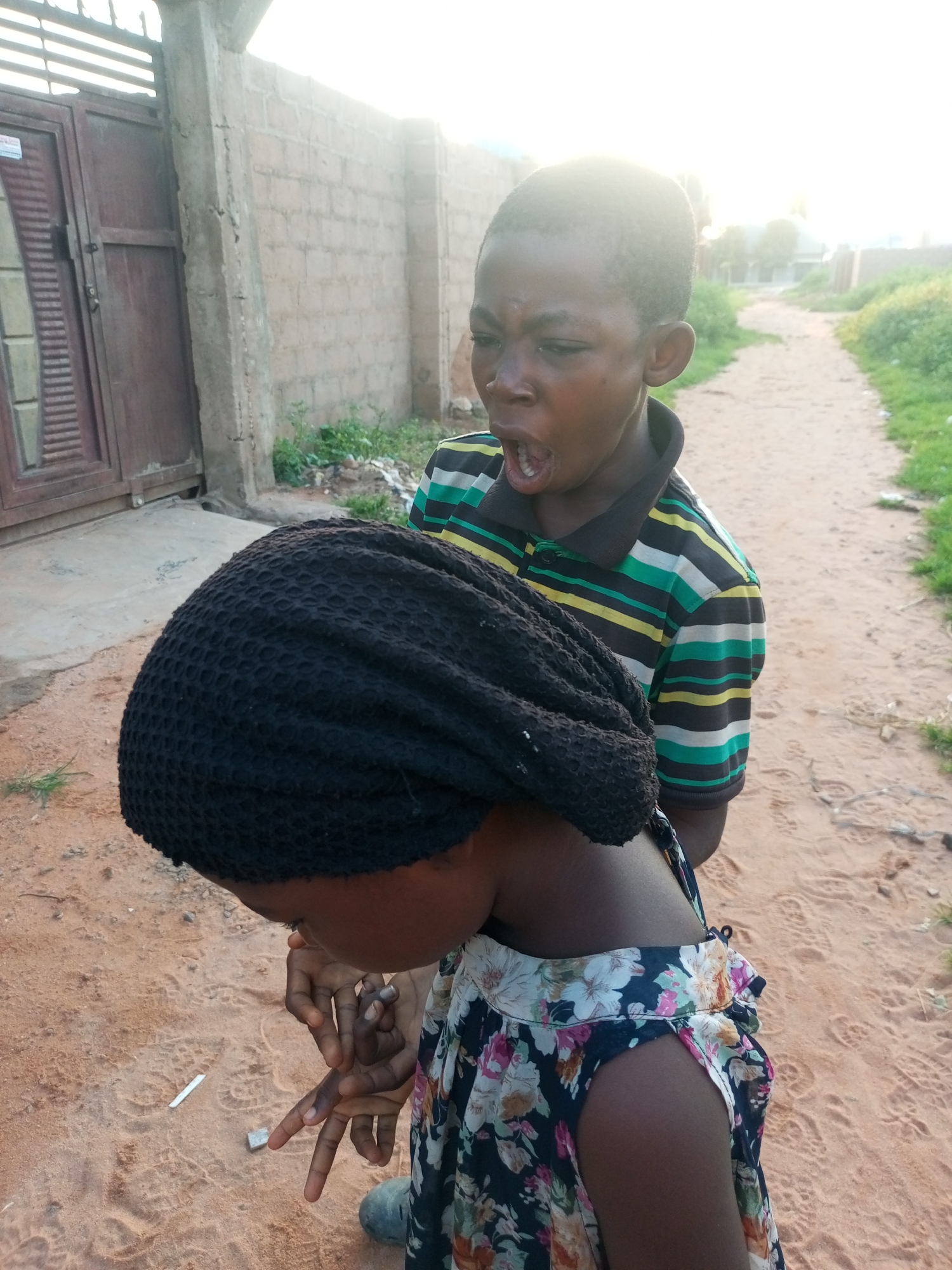 In Tempure, Nigeria, a boy with dark skin and short black hair, wearing a striped shirt with green, yellow, and black bands, stands with his mouth open. He appears to be speaking or shouting. In front of him, a child with dark skin, wearing a black head covering and a floral dress, is looking down at their hands, which are near the ground. The child's hands are positioned as if interacting with something on the dusty, sandy surface. A small white object, possibly a cigarette butt, and a metal object are visible on the ground near the child's hands. The background shows a brick wall, a metal gate, and a dirt path lined with green vegetation. The lighting suggests it is daytime, possibly late afternoon, with the sun creating a bright glare.