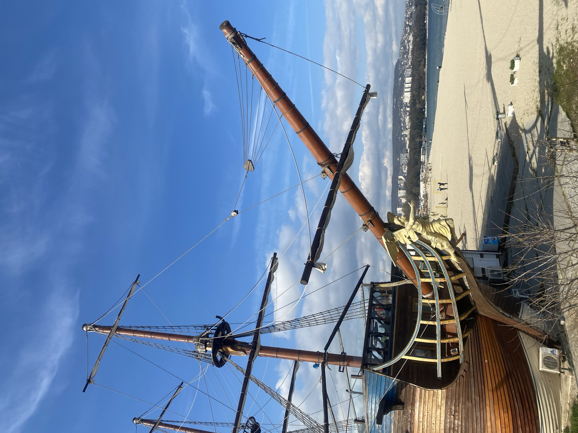 The image shows a partial view of a pirate ship replica located in Varna, Bulgaria. The ship has wooden masts and rigging against a blue sky with scattered clouds. In the background, a beach is visible with people walking along the shoreline. Buildings can be seen further in the background, possibly part of the city's coastal area. The ship's stern is decorated with a figurehead. A modern air conditioning unit is visible on the side of the building.