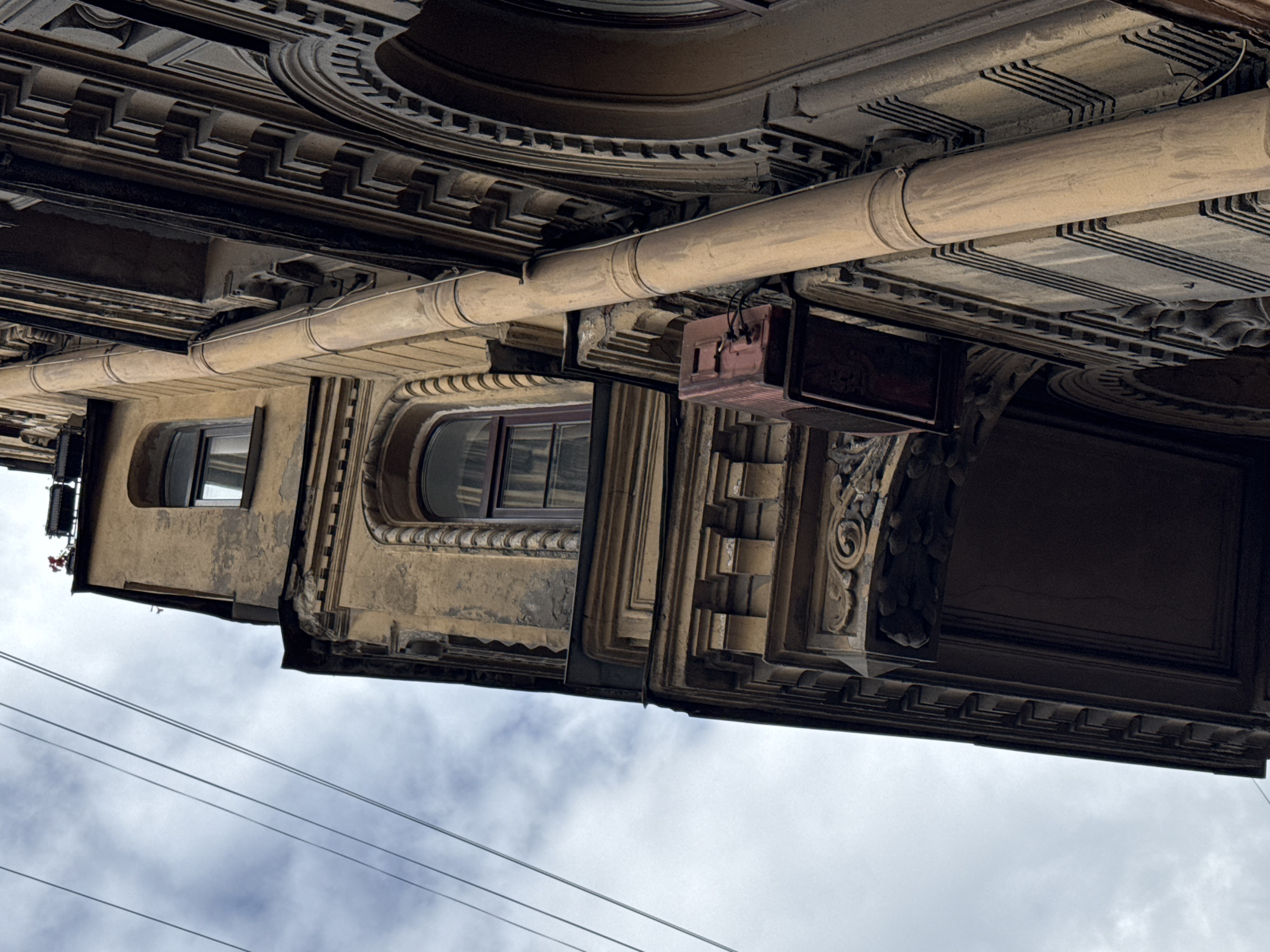 The image is an upward-angle shot of an old, weathered building facade in Saint Petersburg, Russia, under a cloudy sky. The building features ornate architectural details, including decorative cornices, arches, and textured stonework. A large, beige downspout runs vertically along the facade. Attached to the downspout is a red, rectangular electrical box with wires extending from it. Multiple thin, dark electrical wires are also visible in the upper left portion of the frame, running diagonally against the sky. The building's stuccoed walls are a faded yellow-ochre color and show signs of peeling and age. The overall impression is one of historical architecture in an urban setting.