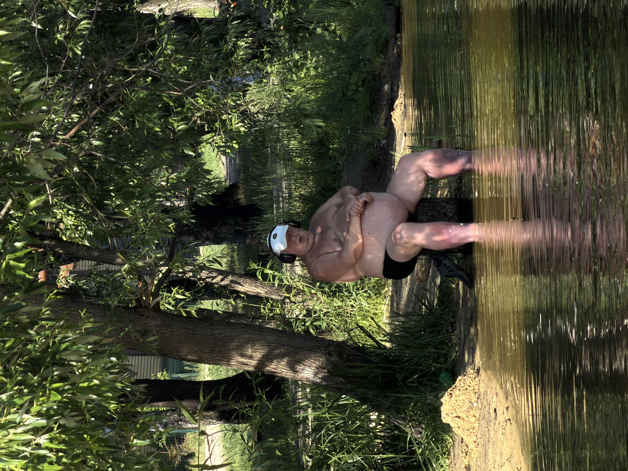 A man with headphones is sitting on a log at the edge of a body of water in Malakhovka, Russia. He is wearing black swim shorts and appears to have his feet in the water. The area is surrounded by trees and lush green vegetation. The water is murky and reflects the surrounding greenery and sunlight. There is a small pile of sand on the bank to the left of the man. The man is looking towards the viewer with his eyes closed and mouth slightly open. The scene appears to be a peaceful outdoor setting.