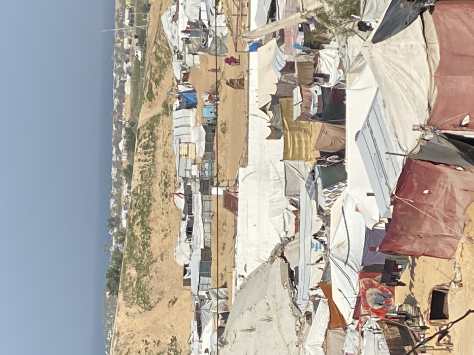 The image displays an elevated view of a densely populated informal settlement located in Khan Yunis, Palestinian Territory, under clear, bright daytime conditions. The scene is dominated by numerous makeshift shelters, tents, and shacks constructed from various materials, including white, beige, blue, and maroon tarpaulins, fabric sheets, corrugated metal, and wood, arranged closely together on sandy terrain. Clothes are visible hanging on lines between several structures in the foreground and midground. A prominent white retaining wall or partially constructed/demolished structure runs horizontally across the midground. Several individuals are present on dirt paths: one figure in a pink garment walks rightward in the upper midground, accompanied by two smaller figures. Additional individuals are visible near blue and white shelters further left in the midground. The background features a large, sandy hillside with sparse green vegetation, beyond which the Mediterranean Sea is visible under a clear blue sky, flanked by a distant strip of coastal buildings and scattered trees. Faint power lines or wires are discernible across the upper midground.