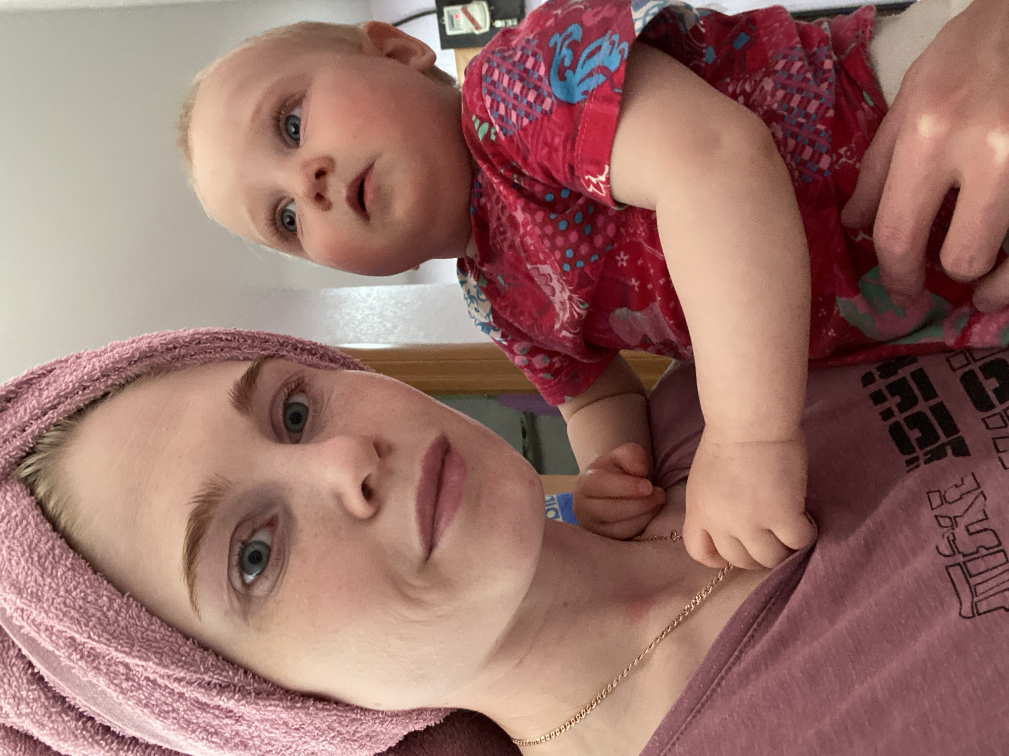A light-skinned woman wearing a pink towel wrapped around her head and a pink t-shirt with black lettering is holding a light-skinned baby. The baby is wearing a red patterned shirt. The baby is looking at the camera. The location is Chaikovsky, Russia.