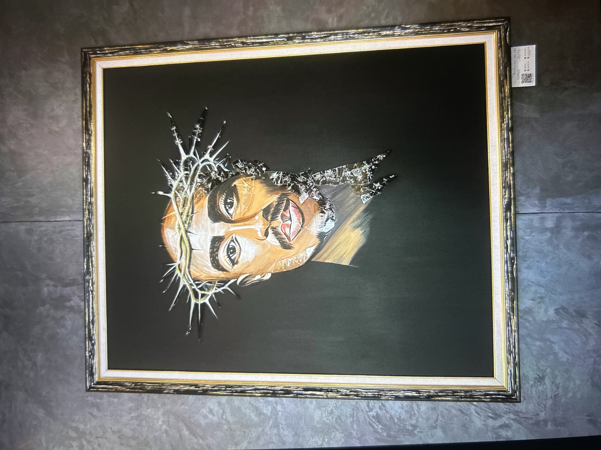 A framed painting hangs on a textured gray wall. The painting depicts the face of a person with dark skin wearing a crown of thorns. The person has a mustache and goatee, and their eyes are looking forward. The right side of their face appears to be made of fragmented, shiny material. The background of the painting is black. A small white tag with QR codes and text is affixed to the bottom right corner of the frame.
