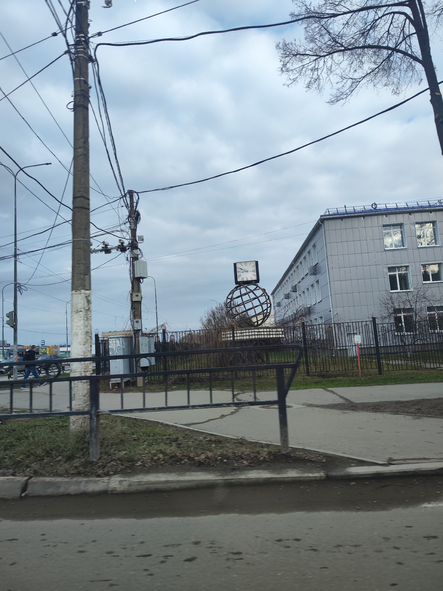 The image shows a street scene in what appears to be Chaikovsky, Russia. In the background is a light gray, multi-story building with rows of windows. A decorative metal globe with a clock on top stands behind a black metal fence, near a sidewalk. An electrical pole with multiple wires is located on the left. A person is walking across the street in the background. A puddle is visible on the road in the foreground. The sky is overcast.