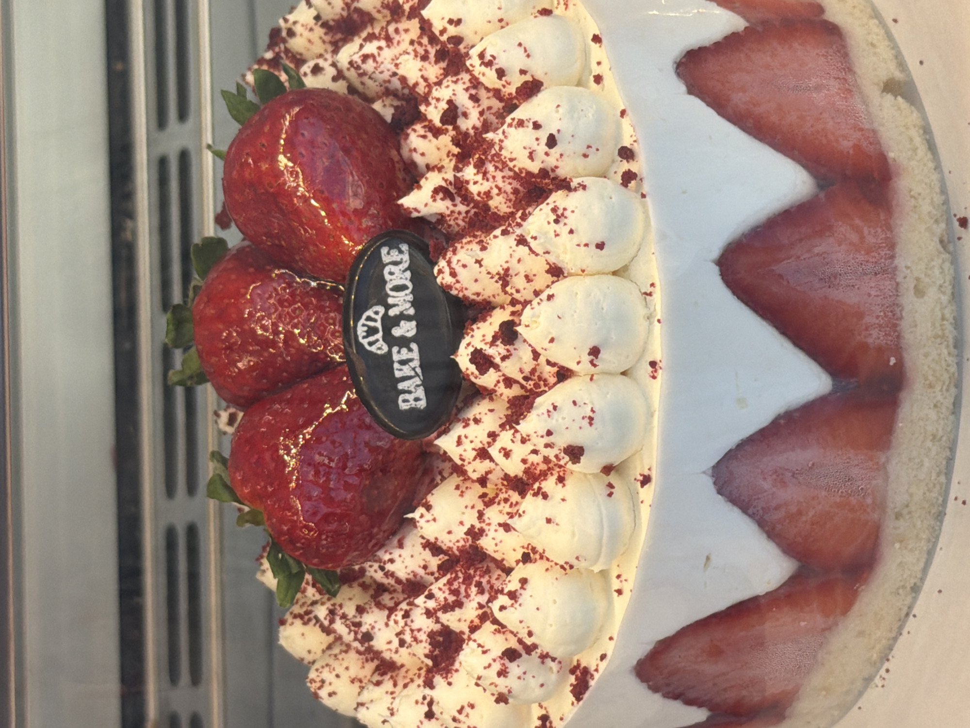 This vertically oriented close-up image showcases a meticulously decorated round cake, likely displayed in a refrigerated case. The top of the cake is richly adorned with several whole, glossy red strawberries and generous dollops of white cream or frosting, which are generously sprinkled with small red crumbs, possibly from red velvet cake. Nestled among the strawberries and cream is a small, oval-shaped black plaque featuring a white croissant icon above the words "BAKE & MORE" in white capital letters.
The side of the cake reveals a distinctive design where vibrant red strawberry slices are artfully embedded within the white cream layer, forming a wavy, chevron-like pattern. This side cream layer appears to sit on a light-colored cake base. Some of the strawberries on the side exhibit slight condensation, reinforcing the impression of a refrigerated display.
The background to the left is blurred, showing vertical grey or metallic bars, consistent with the interior of a display cabinet or a shelving unit. The image focuses entirely on the cake, with no people or specific activities taking place. The overall impression is one of an appealing dessert, professionally presented for sale or display in a bakery or cafe setting, potentially in Fethiye, Turkey.