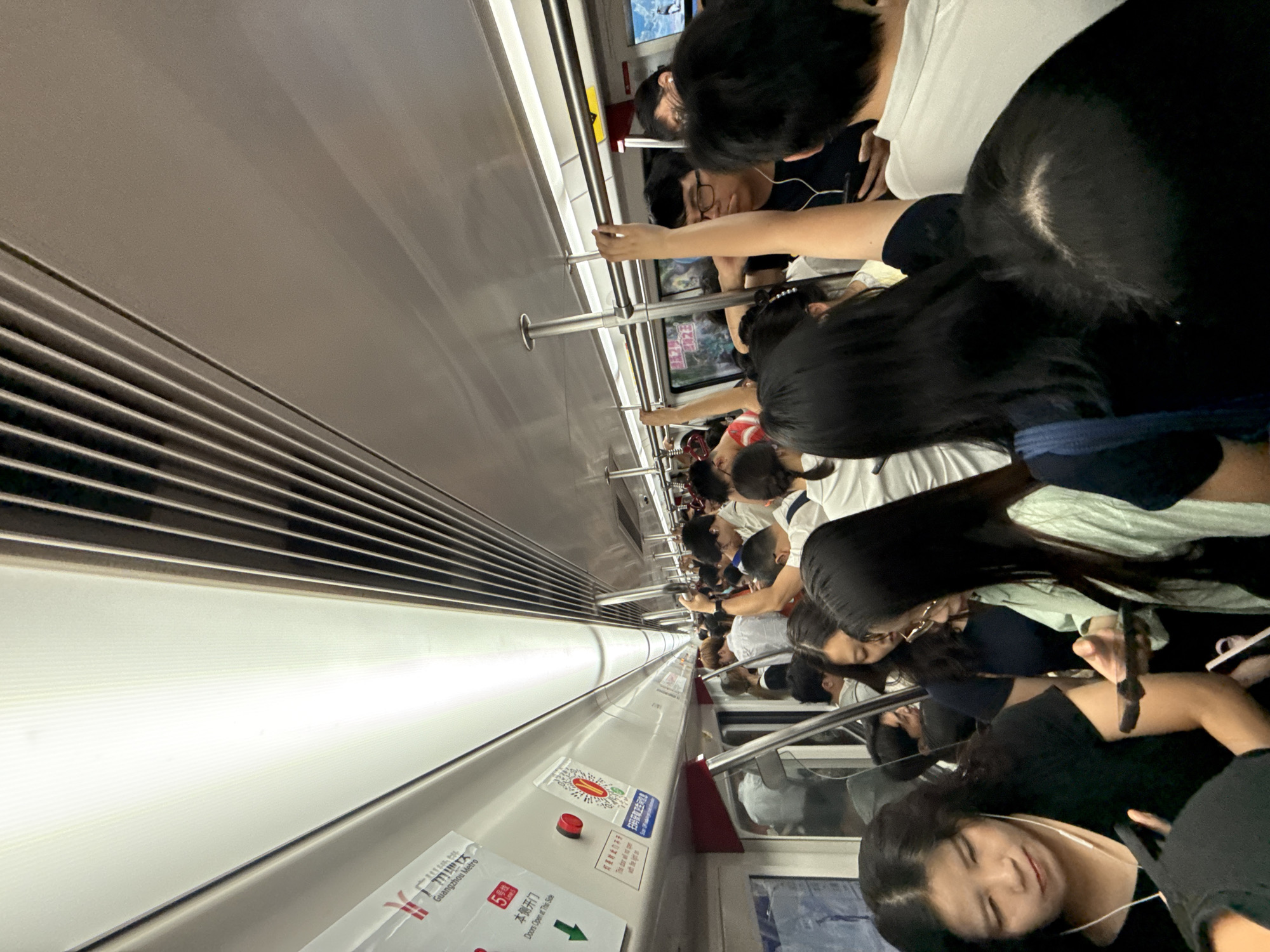 The image depicts a crowded interior of a subway train, likely in Guangzhou Metro, Tianhe District, China, given the signage that reads "Guangzhou Metro." The scene is characterized by a high density of passengers standing, with limited space to move. Various individuals are visible, some holding onto overhead bars for support, while others are in conversation or looking at their mobile phones. The train car features a white interior with ribbed ceiling panels and bright fluorescent lighting. Signs with Chinese characters and directional arrows are affixed to the walls. The overall impression is one of a busy commute during peak hours.