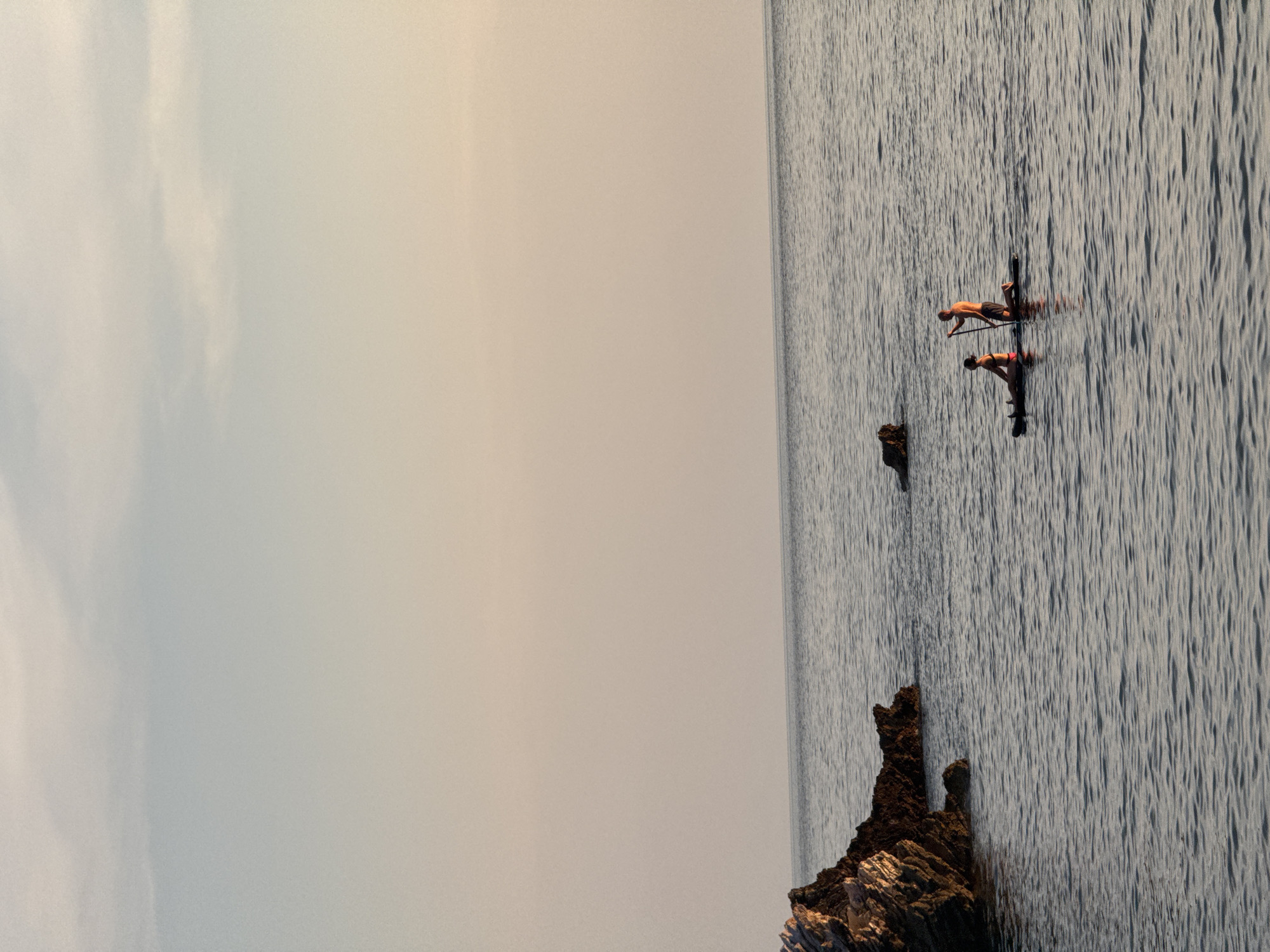 In Sveti Stefan, Montenegro, two individuals are paddleboarding on the calm sea during sunset. A woman, wearing a bikini, is seated on the paddleboard. A man, shirtless and wearing shorts, is kneeling behind her and paddling. The sun is low in the sky, casting a warm glow on the scene. Rocks are visible on the left side of the image, emerging from the water. The horizon line is sharp, separating the sea from the soft hues of the sky.