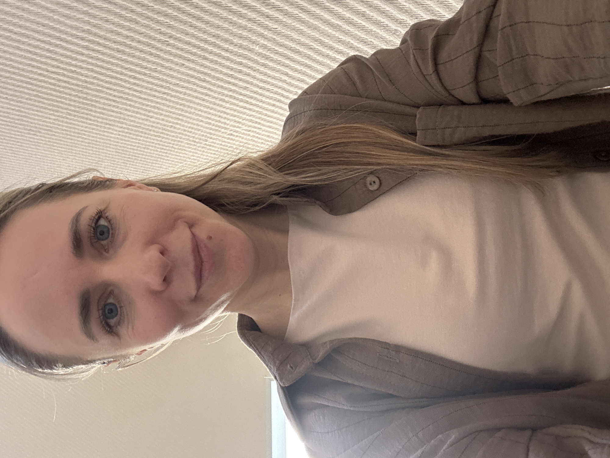 A woman with blonde hair and blue eyes is looking down at the camera. She is wearing a white t-shirt and a light brown button-down shirt. The background appears to be a ceiling with a textured surface. The location is not identifiable from the image.