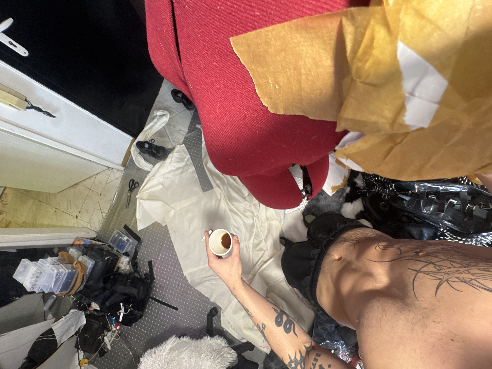 The image depicts an indoor scene with a person holding a small cup of dark liquid. The person's arm, covered in tattoos, is visible. They are wearing black shorts and a pierced navel is visible. A pile of translucent plastic containers with various items inside is stacked in the background, along with a black backpack and other bags. A white cloth is spread on the floor, and a red textile is visible in the foreground. The floor appears to be tiled and metal. The scene does not provide any specific indicators to determine the location within Sofia, Bulgaria.