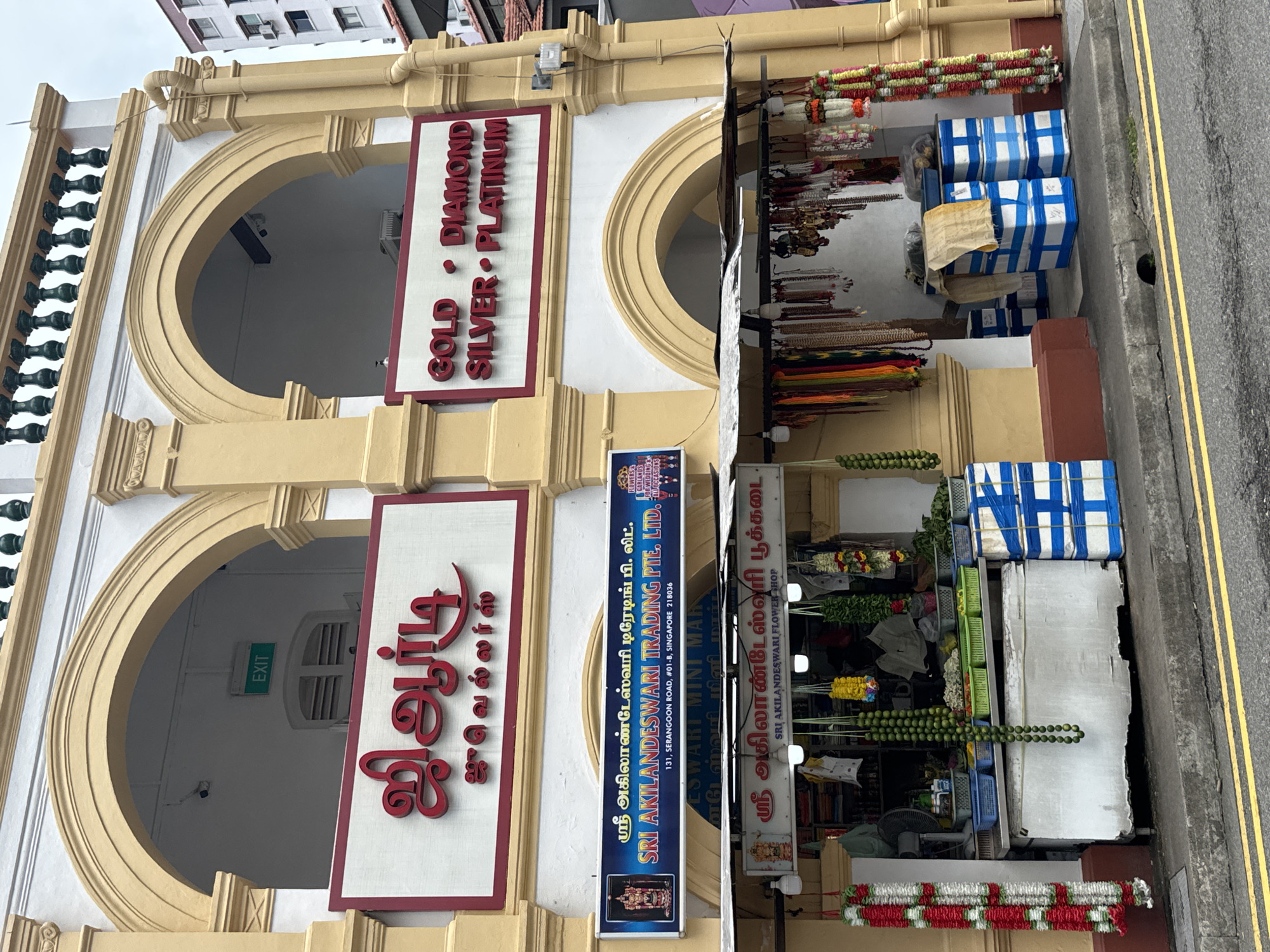 The image shows a shopfront in Little India, Singapore. The shop is a jewelry store and a flower shop. The jewelry store has a sign in Tamil that says "Sri Akilandswari Trading Pte. Ltd." The flower shop has a sign that says "Sri Akilandswari Flower Shop." The shop is decorated with garlands of flowers and beads. There are also some people in the shop, but they are not clearly visible. The shop is located on Serangoon Road, which is a busy street in Little India.